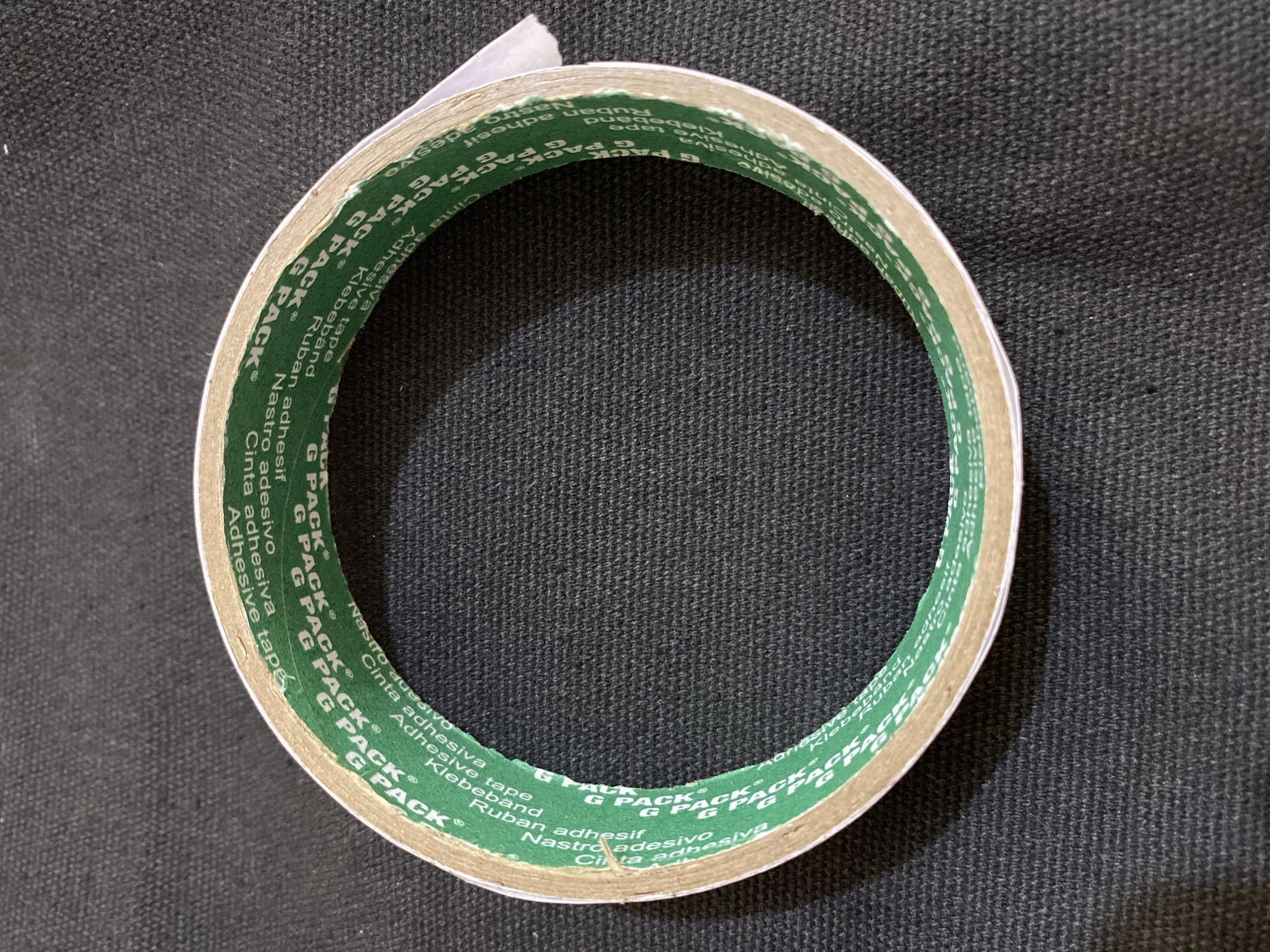 The image displays a roll of green and white adhesive tape positioned on a dark, textured surface. The tape is circular and appears to be made of two layers with adhesive on both sides. The green portion of the tape is printed with repeated instances of "G PACK" and various other words in different languages, including "Adhesive Tape," "Cinta Adhesiva," and "Ruban Adhésif," suggesting it is a multi-purpose adhesive product. The white layer is the backing paper, partially peeled back at the top right of the roll. The background surface is consistent with a dark fabric or carpet. There are no people or signs of specific urban location details in the image. The overall impression is a close-up product shot.