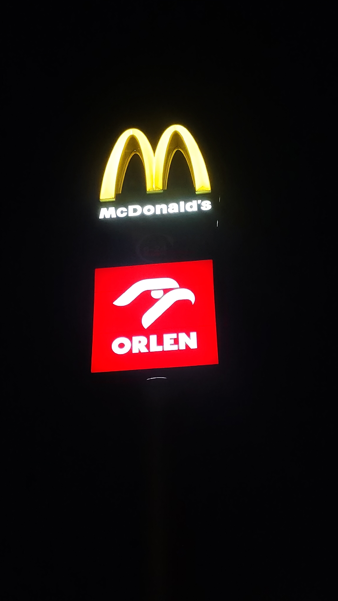 The image shows two signs against a black background. The top sign features the golden "M" logo of McDonald's, with the text "McDonald's" in white lettering below it. The bottom sign is a red square with a white logo that appears to be an eagle or bird of prey, with the word "ORLEN" in white capital letters below the logo. The McDonald's sign is positioned above the Orlen sign. The image appears to be taken at night, as indicated by the dark background and the illuminated signs. The setting is a city location, specifically Rudná, Czechia, likely a commercial area with these establishments. There are no people or other visible objects or activities in the scene.