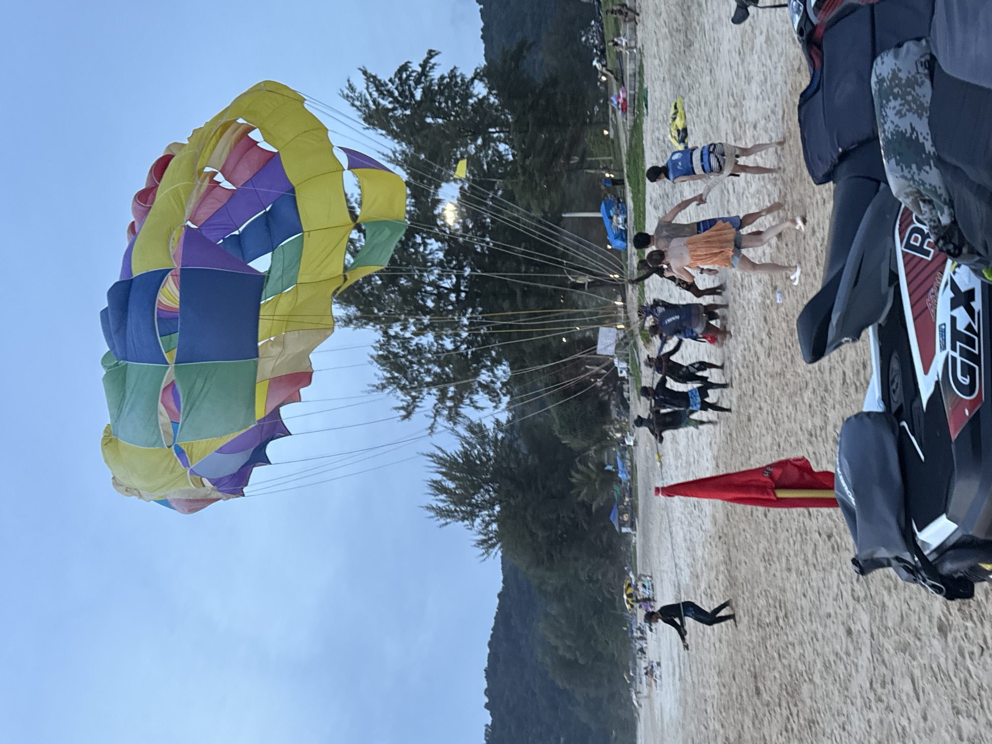 The image depicts a parasailing activity on Karon Beach, Thailand, during dusk. A large, colorful parasail, a canopy of yellow, green, blue, and purple panels, is visible in the sky. Several people are gathered on the sandy beach, some appearing to be involved in preparing or operating the parasailing equipment. Two individuals, possibly participants, are being pulled by ropes attached to the parasail, with one wearing an orange top and denim shorts, and another a blue life vest. A group of at least four people in dark attire, likely staff, are near them, holding onto ropes. A bright red flag on a pole stands prominently on the sand in the foreground. A jet ski is partially visible in the bottom right corner of the image, with some gear on it. In the background, the beach stretches out with scattered beach umbrellas and loungers, and a line of lush green trees and hills forms the backdrop. The sky is overcast, indicating either early morning or late evening.