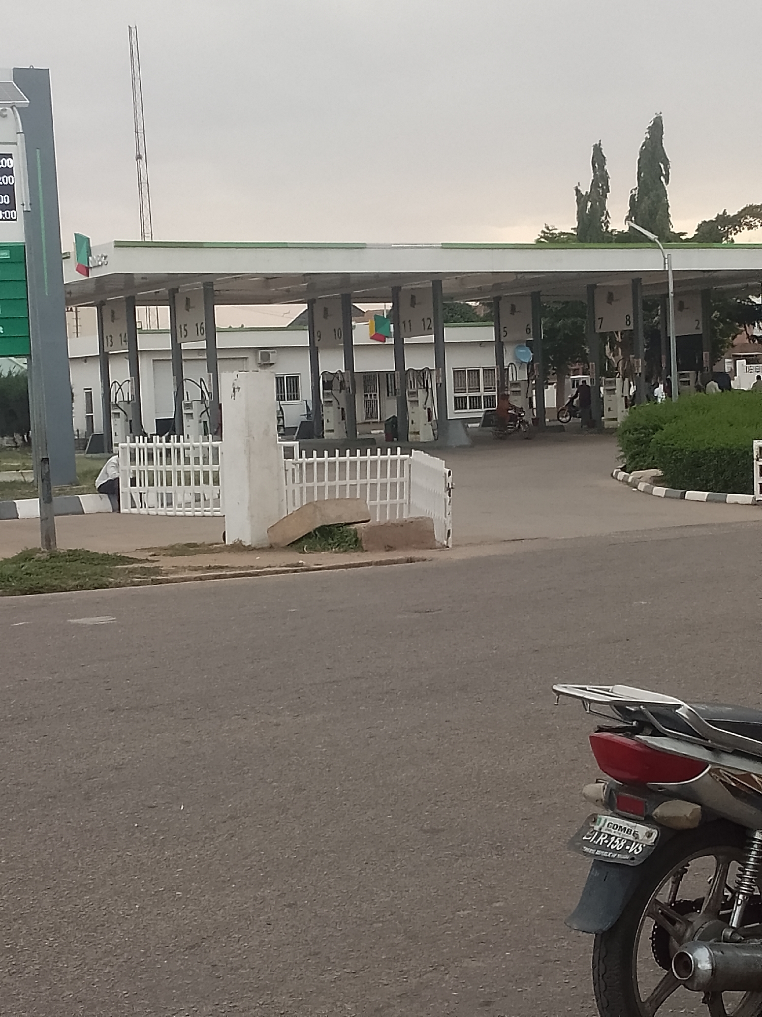 The image shows a gas station in Pantame, Nigeria. A white motorcycle is parked in the foreground, with its license plate visible. In the background, a series of gas pumps are lined up under a covered structure. The sky is overcast, and the scene appears to be outdoors. There are a few people visible in the background, some of whom appear to be employees of the gas station. The overall impression is one of a typical day at a gas station in Nigeria.