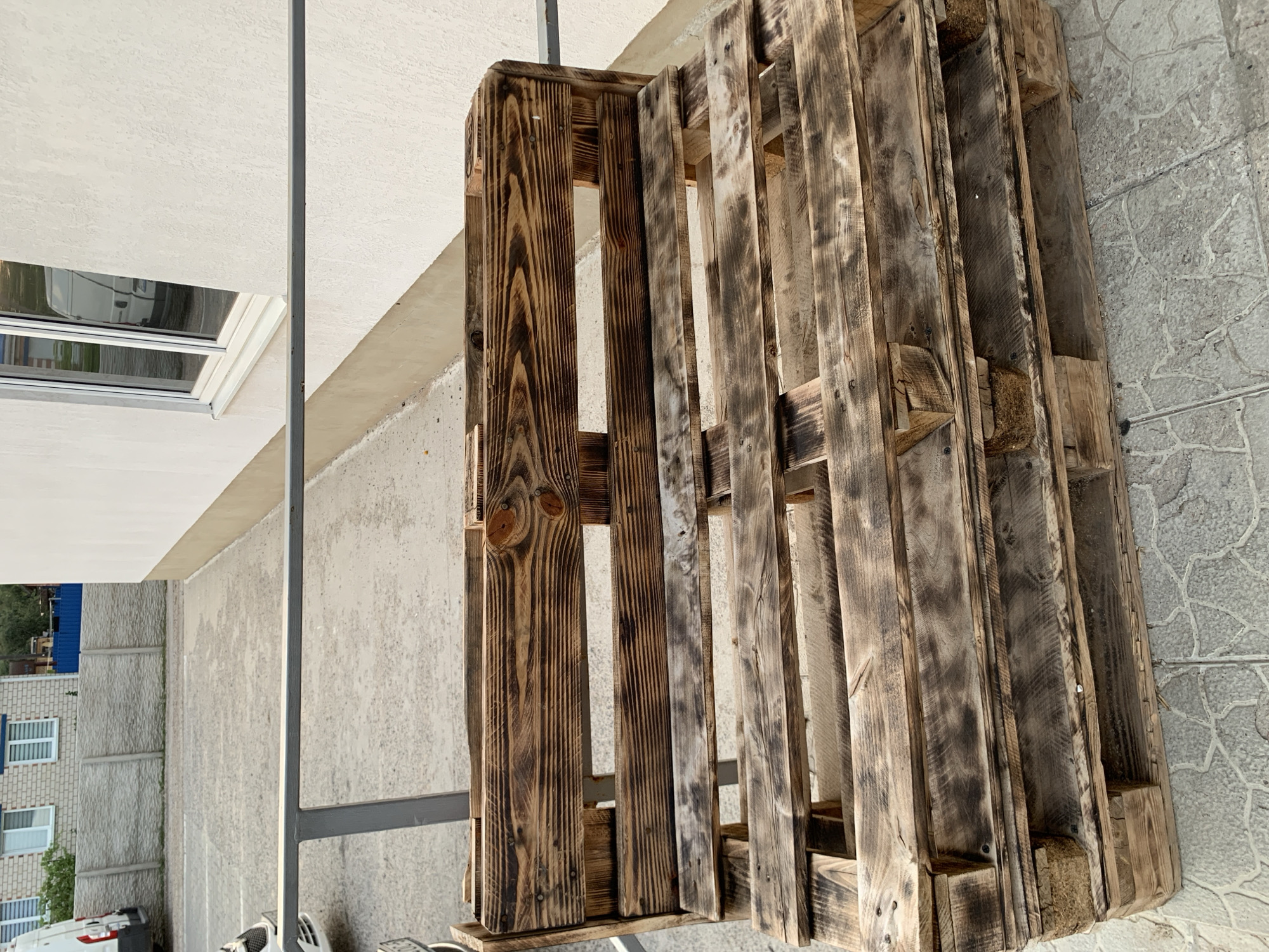 An outdoor scene captures a rustic bench constructed from wooden pallets. The bench is positioned on a grey paved surface, with a light-colored building and a grey concrete fence visible in the background. A metal railing runs horizontally across the middle ground of the image. The bench itself shows signs of weathering and charring on the wood, suggesting it has been exposed to the elements and possibly treated with heat. The overall environment appears to be a utilitarian or commercial space, likely a yard or an outdoor seating area associated with a building. The location is identified as Kapyĺ, Belarus. There are no people or animals visible in the image.