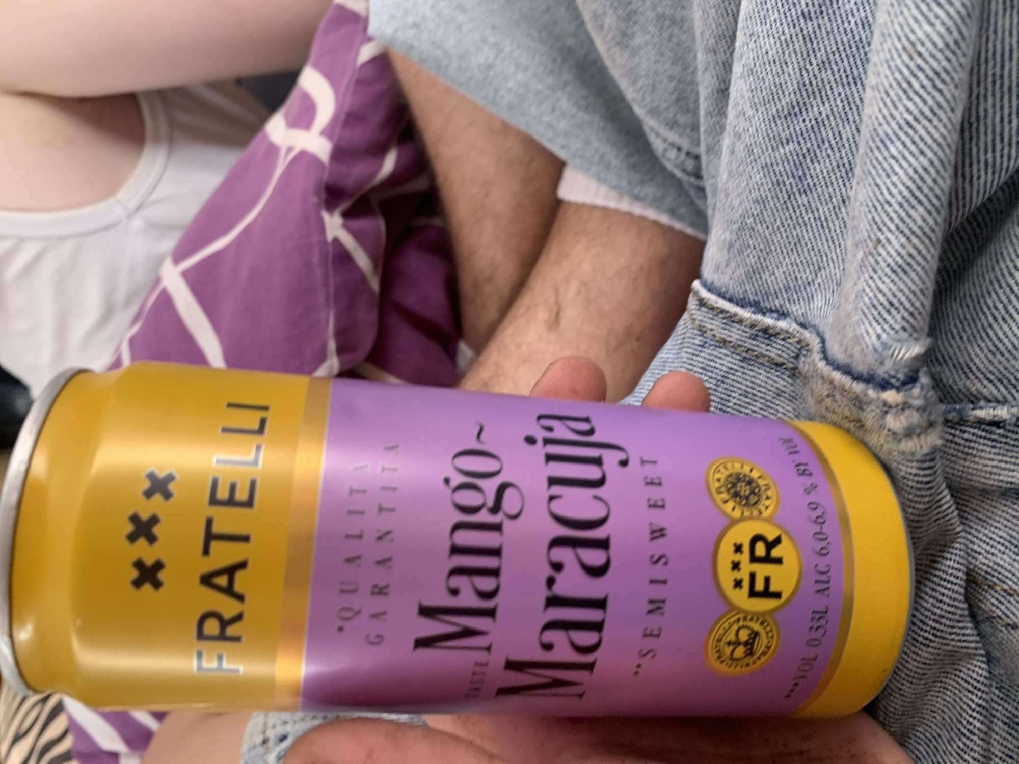 A person is holding a yellow and purple can of "FRATELLI Mango-Maracuja" in their hand. The can has text indicating "QUALITA GARANTITA" and "SEMISWEET". Below this, there are circular emblems, one with a crown, another with "FR" and three crosses, and a third with a decorative pattern. At the bottom of the can, there is text stating "VOL 0,33L ALC 6,0-6,9 % BY VOL". The person is wearing denim jeans, and a purple patterned blanket and a white tank top are visible in the background, suggesting an indoor or relaxed setting. The image does not contain any information that can be used to determine the location within the city of Zhmerynka, Ukraine.