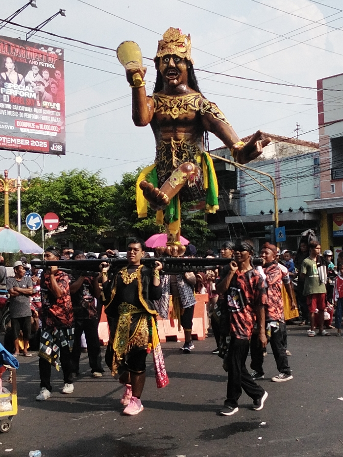 A daytime street scene in Probolinggo, Indonesia, depicts a cultural procession. The central focus is a large, elaborate float portraying a dark-skinned male figure with a fierce expression, golden crown, intricate gold-patterned attire, and vibrant yellow and green fabric adornments. The figure holds a golden goblet-like object in its raised right hand and has its left hand extended.
Below the float, a group of approximately six men are carrying a dark, thick horizontal pole supporting the structure. One man in the foreground, center-left, wears a traditional black and gold garment with bright pink sneakers and glasses. Other carriers wear casual attire, including patterned t-shirts and dark pants. A pink umbrella is visible directly beneath the float.
In the background, a large red and black banner advertises an "ALL CAR MEET UP" event scheduled for "SEPTEMBER 2025" in Probolinggo. Street signs visible include a blue circular sign with a white left arrow and a red circular "No Entry" sign. Buildings, power lines, and green trees line the street. Numerous other individuals are visible in the background, observing or participating in the procession. The asphalt road appears partially damp. The scene is brightly lit by natural sunlight, casting visible shadows.