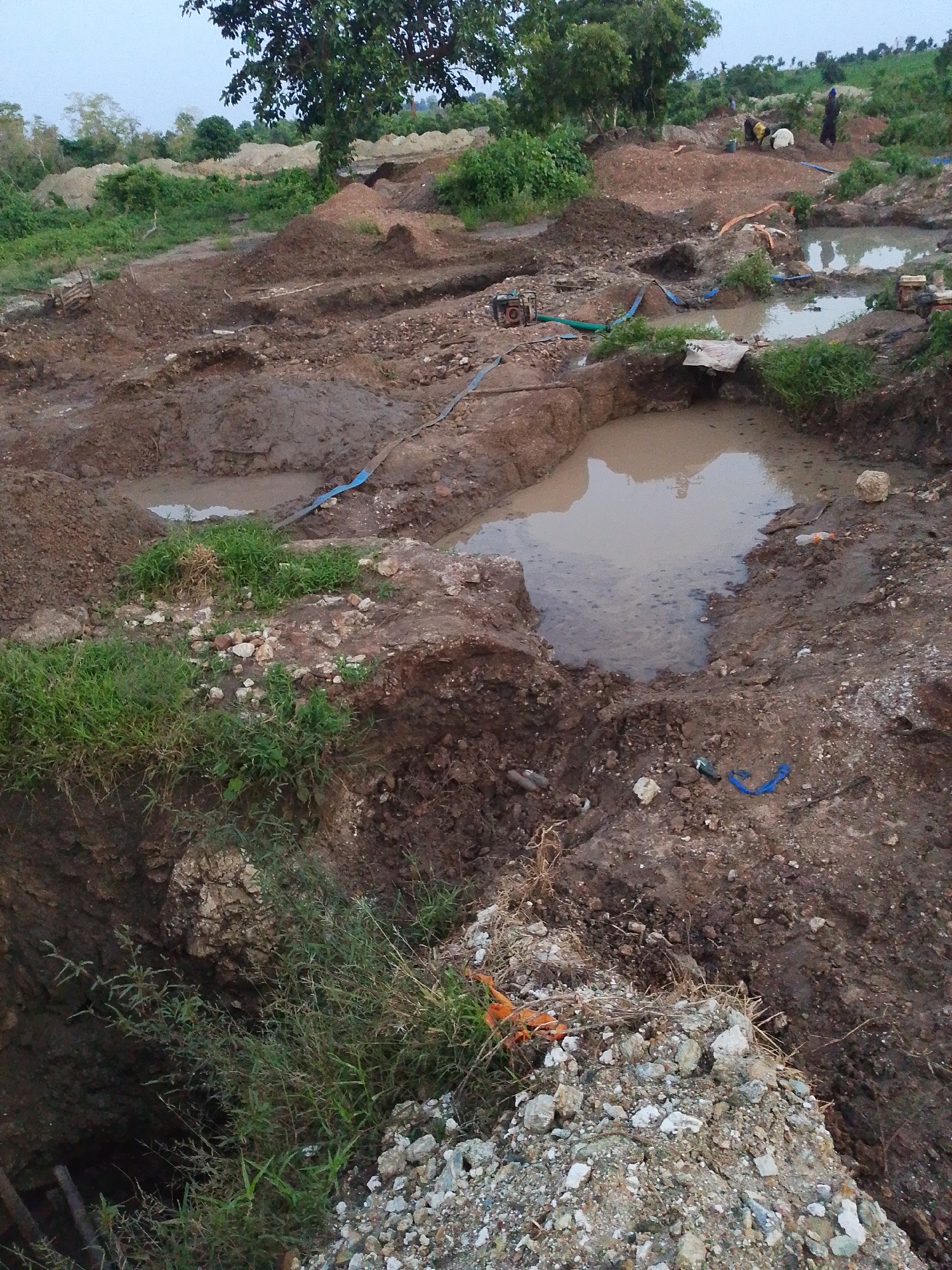 The image depicts a landscape in Otulu, Nigeria, characterized by a disturbed terrain suggestive of excavation or mining operations. Several pools of murky water are present, interspersed with mounds of excavated earth and debris. Patches of green vegetation, including grass and trees, are visible, contrasting with the predominantly brown and reddish hues of the soil.
In the mid-ground, three individuals are present. One person, wearing dark clothing, stands near a pool of water. Another individual, also in dark attire, appears to be working near a pile of sand. A third person, dressed in yellow and white, is also engaged in activity in the vicinity. Various objects, including blue hoses, what appear to be pipes, and possibly equipment for pumping or sifting materials, are scattered throughout the scene. The overall impression is one of an active, though rudimentary, industrial or agricultural site. The sky is overcast, suggesting potentially subdued lighting conditions.
