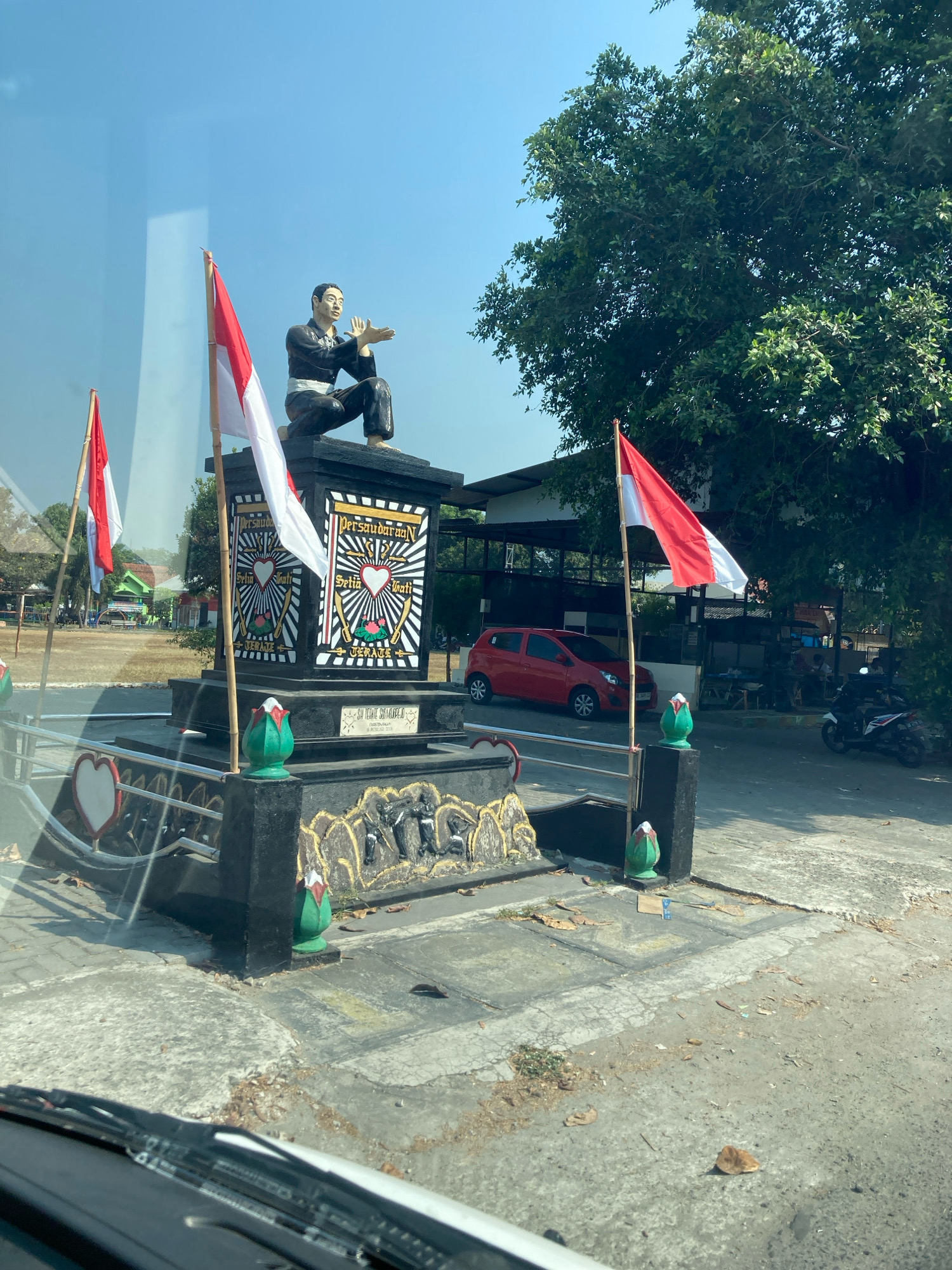 A statue of a man in a martial arts pose is displayed on a monument. The monument is decorated with Indonesian flags, which are red and white. The statue is kneeling and has his hands raised in front of him. There are also inscriptions on the monument. In the background, there are trees, a red car, and a motorcycle. The scene appears to be outdoors in a public space.