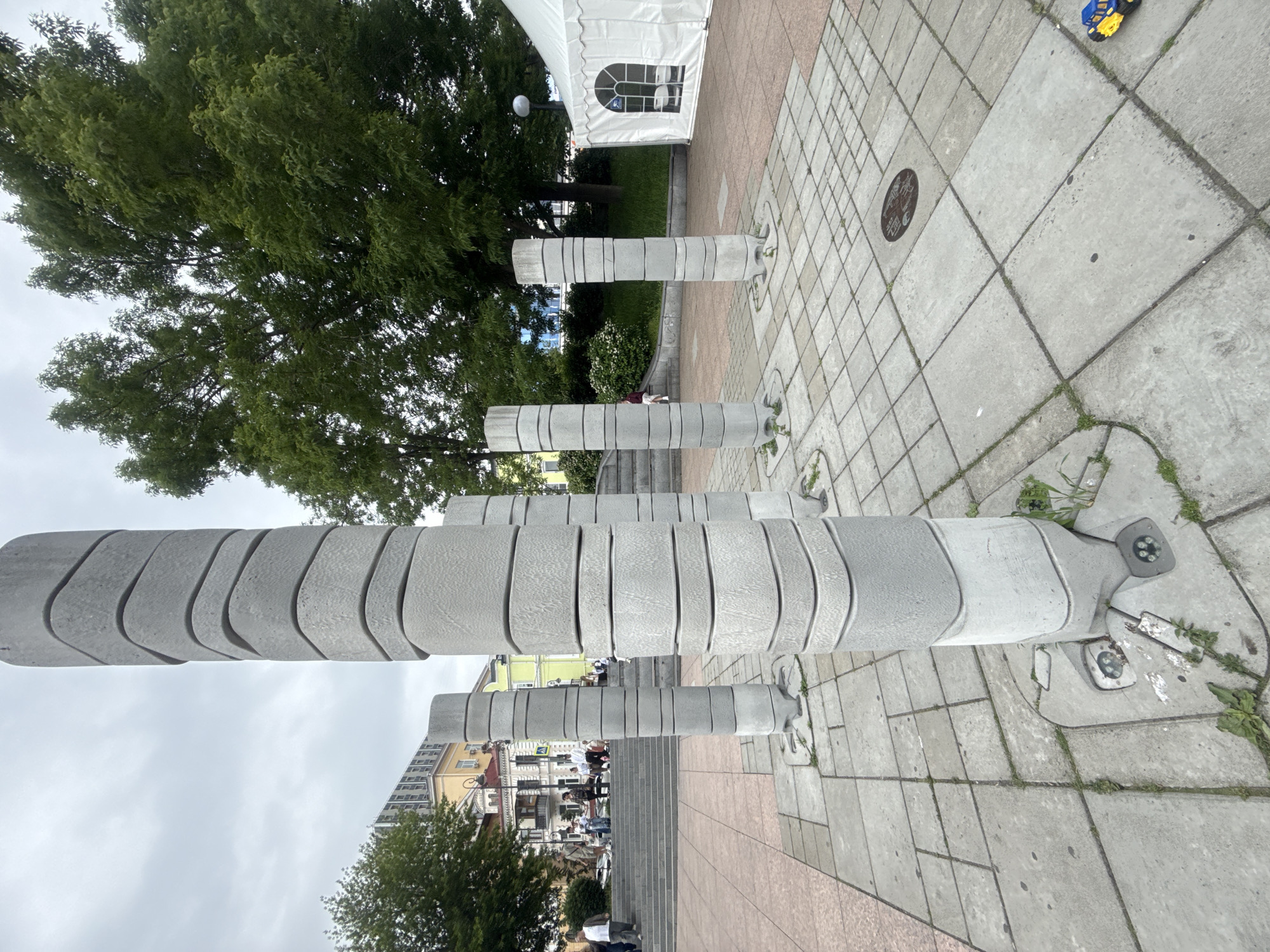 The image shows an outdoor public space in Vladivostok, Russia, featuring a cluster of abstract concrete pillars arranged on a paved plaza. The pillars are tall and cylindrical, constructed from stacked, curved concrete segments, giving them a layered appearance. They are illuminated from below by integrated lighting fixtures. The plaza is paved with large, square concrete tiles.
In the background, there are several buildings with varying architectural styles, some appearing older with ornate facades, and others more modern. Green trees with dense foliage are visible, providing shade and contrast to the concrete structures. A white tent structure, possibly for an event or market, is situated to the right of the frame.
Several individuals are present in the scene. On a raised set of stairs to the left, a group of people are walking and standing, some appearing to be tourists or locals enjoying the public space. In the foreground, a small toy car, yellow and blue, is placed on the pavement. The sky is overcast, suggesting a cloudy day. The overall impression is of a city square with artistic installations and surrounding urban infrastructure.
