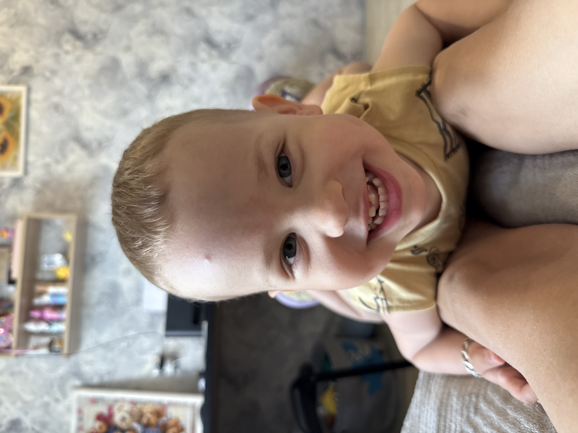 A young child with blond hair and blue eyes is smiling broadly, revealing his teeth. He is wearing a yellow shirt. The child is being held, and an adult hand with a silver ring is visible. The background is blurred but appears to be indoors, with a grey patterned wall and furniture. There is a display shelf containing small toys and items. A framed picture can also be seen on the wall. The location is unspecified but has no association with the city of Rusanovo, Russia.