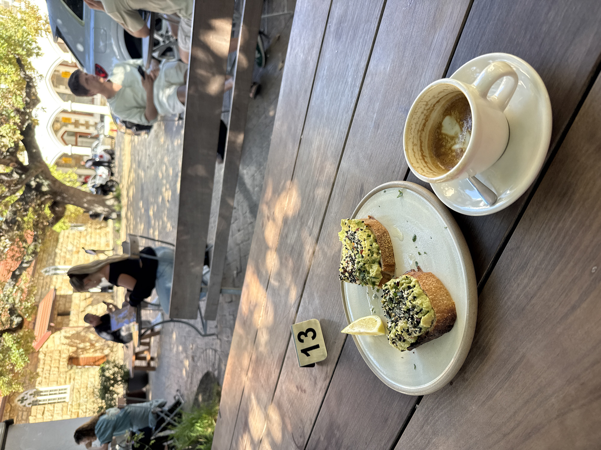 The image shows a cafe scene, likely in Cape Town, South Africa, based on architectural styles. On a wooden table, there's a plate with two slices of avocado toast, sprinkled with seeds and garnished with lemon, and a cup of coffee with a spoon on its saucer. Next to the plate there is a small sign with the number 13. The table is part of a multi-level outdoor seating area. Several people can be seen in the background seated at tables and walking around. The building in the background appears to be a historic structure, with arched windows and stonework. The sky is clear, indicating a sunny day.