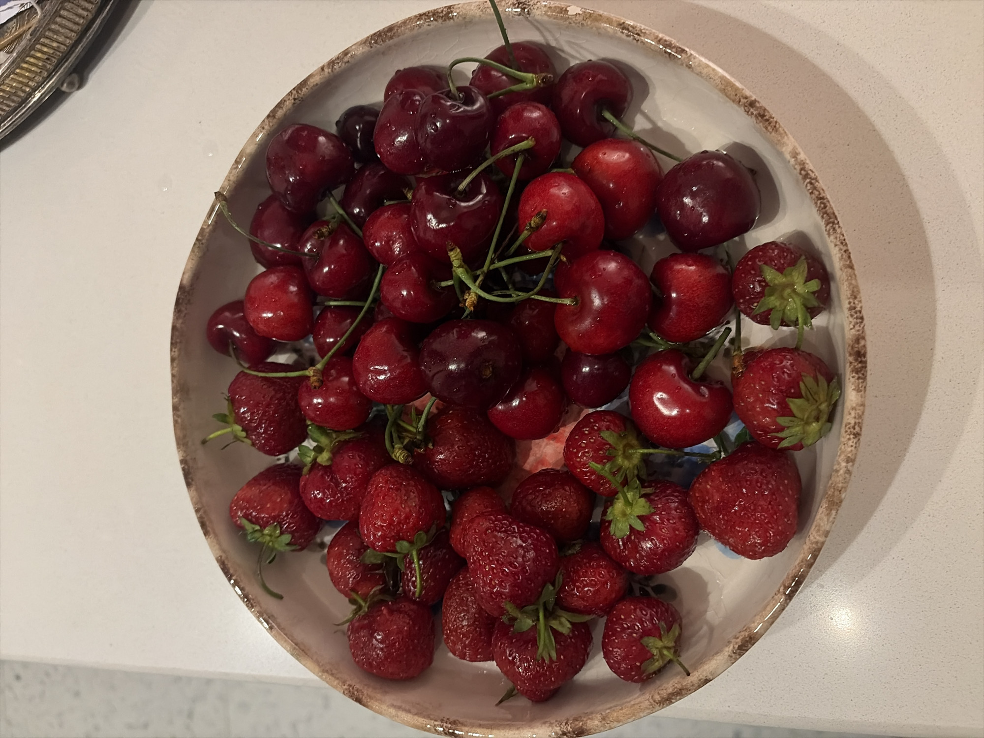 A close-up, overhead shot shows a bowl filled with fresh strawberries and cherries. The bowl is rustic, with a textured, speckled rim. The fruits are vibrant red, some with stems still attached. The background is a plain, light-colored surface, possibly a countertop. There is no indication of the city of Tbilisi, Georgia.
