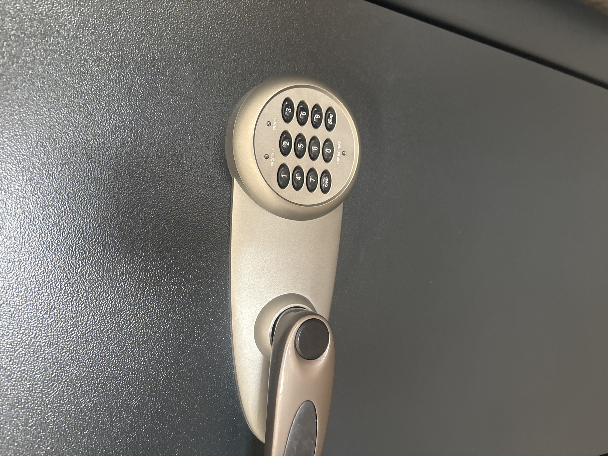 A close-up, eye-level, outdoor shot shows the keypad and handle of a safe. The safe is dark gray with a textured surface. A metallic gold plate surrounds the keypad and handle. The keypad has ten circular buttons numbered 0 through 9, along with buttons labeled "clear", "prog", "PROCEED", and "ERROR". A small light is positioned above the "LOW BATTERY" indicator. The handle is also metallic gold with a dark gray grip. The image shows no people or Dubai-specific landmarks.