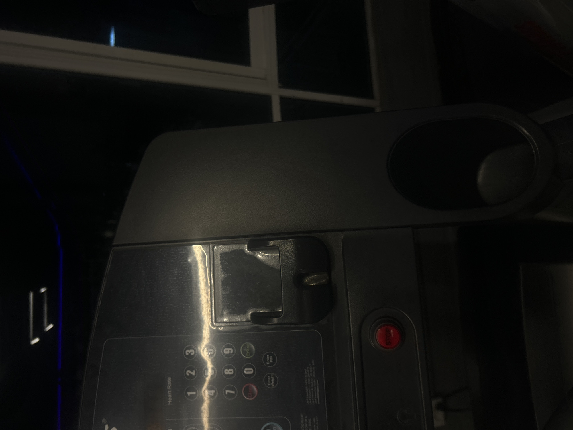 A close-up shot focuses on the control panel of a piece of exercise equipment, likely a treadmill. The panel is predominantly dark gray and features a numeric keypad with buttons labeled "1" through "9" and "0", along with additional buttons marked "Enter PIN", "Enter Weight", and "Clear". A red "STOP" button is visible on the right side of the panel. The words "Heart Rate" are printed above the numeric keypad. The equipment also has a cup holder on the top right. A safety key is inserted in a slot on the upper part of the control panel. In the background, a window with a white frame is partially visible, suggesting the equipment is indoors. The lighting is subdued, creating shadows and reflections on the panel's surface.