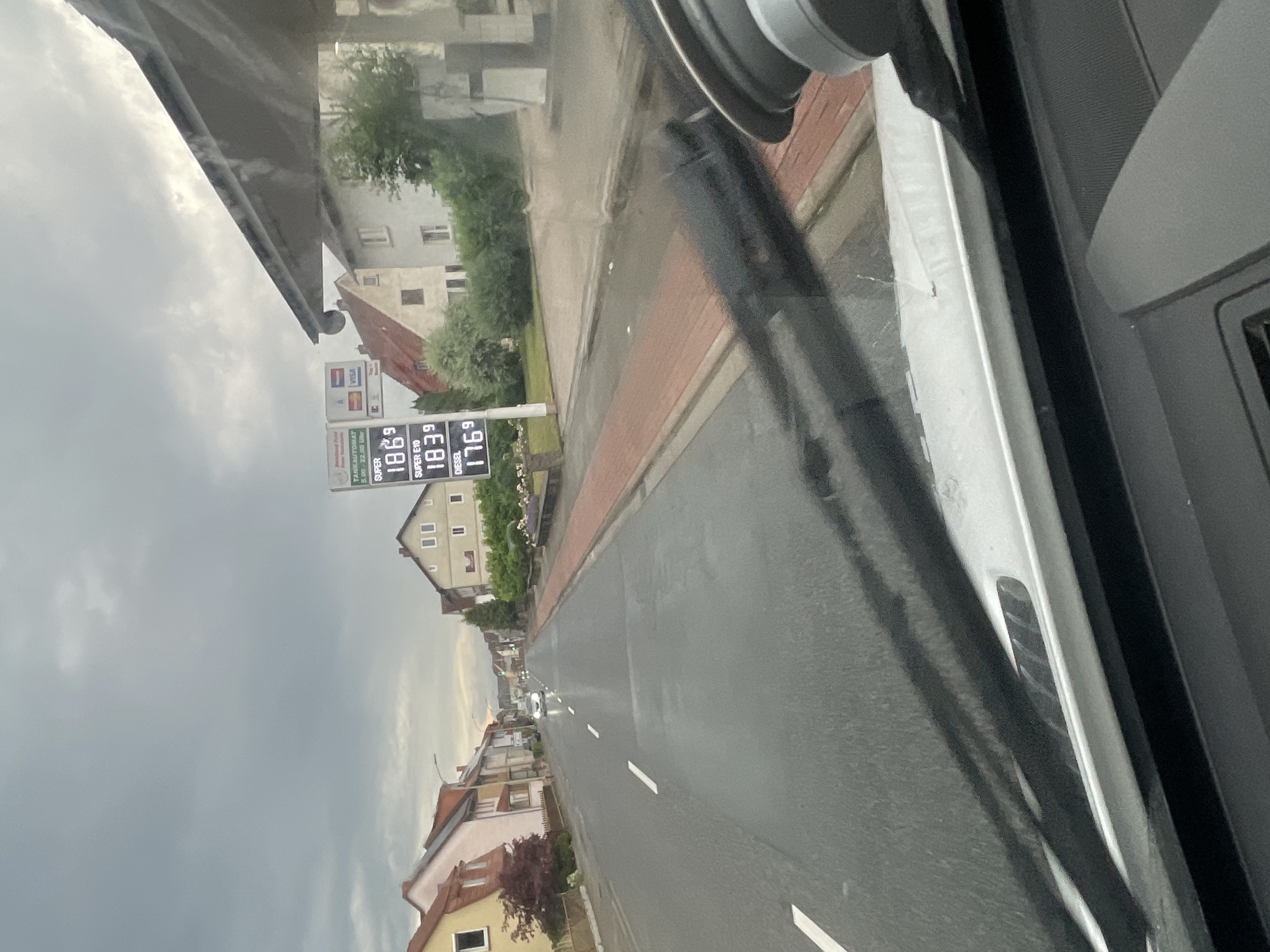 The image shows a street scene in Wartenberg, Germany. The view is from inside a vehicle, looking forward through the windshield and wipers. On the right side of the image is a gas station with a price sign displaying fuel costs. The sign indicates "SUPER 1.86", "SUPER E10 1.83", and "DIESEL 1.76". Houses line the street on both sides. The sky is cloudy and appears overcast. A white car is visible driving down the road in the distance. The road is wet, suggesting recent rain.