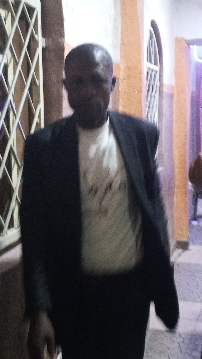 The image depicts an adult male in Sabon Gari East, Nigeria, captured in mid-stride within an indoor or sheltered environment. The subject, appearing to be middle-aged, possesses dark skin and a receding hairline. He is dressed in a dark blazer over a light-colored t-shirt featuring an indiscernible dark graphic design on the chest, paired with dark trousers. His right hand is visible towards the lower left, appearing to hold a small, light-colored object.
The background to the left features a vertical, light-colored metal lattice structure, possibly a gate or window grille. Directly behind the subject, an orange-painted wall segment or pillar includes an arched opening fitted with a similar light-colored lattice grille. Further to the right in the background, a wall section exhibits a purple tint, and a heavily blurred, partially visible figure in a brown garment is present. The floor visible towards the bottom right displays a checkered pattern of light and dark tiles. The scene is illuminated by dim, artificial lighting.