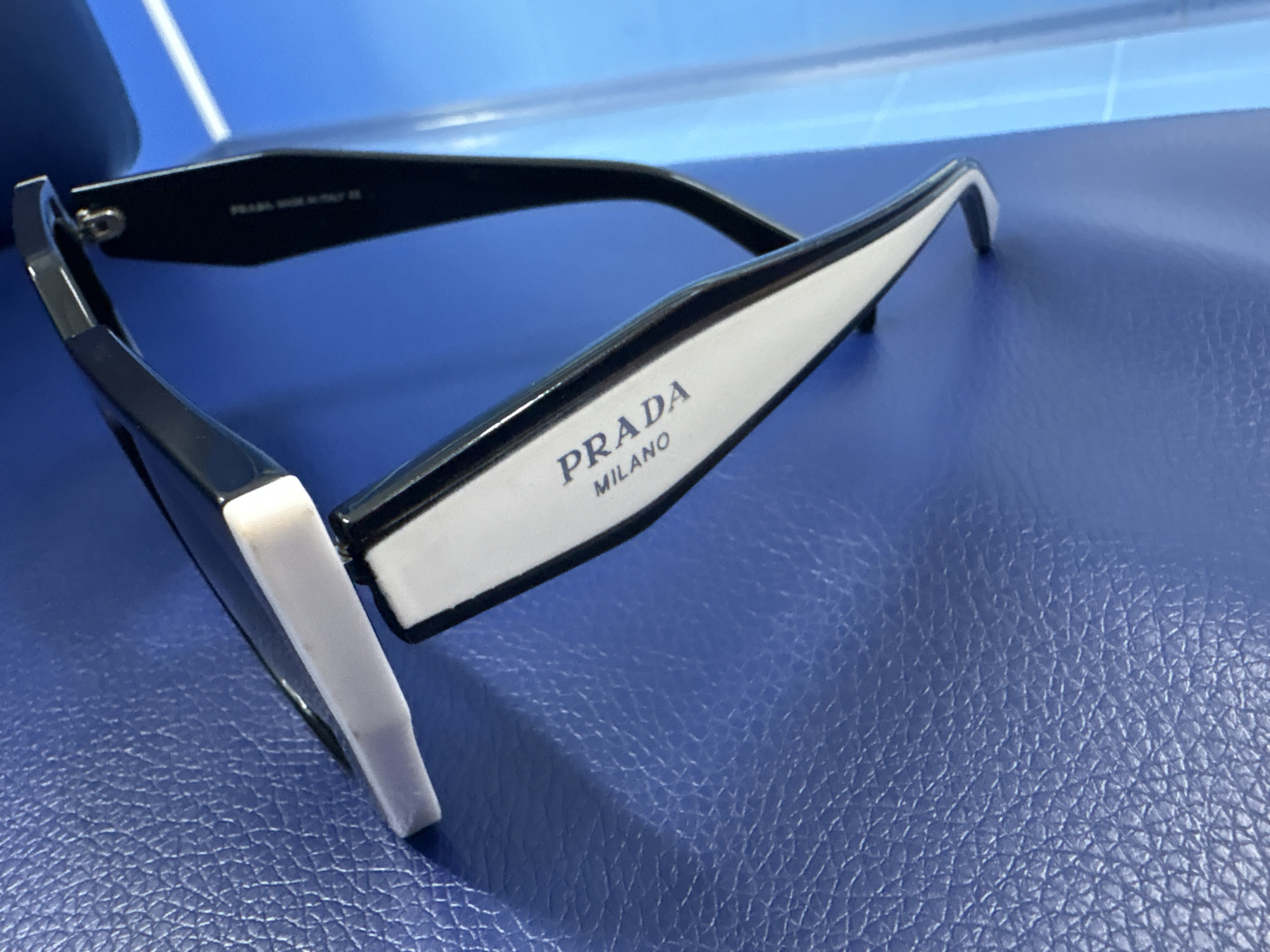 The image shows a close-up, angled view of a pair of black and white Prada sunglasses resting on a textured blue surface. The sunglasses are positioned with the temples folded in and are angled so that the right temple, which features "PRADA MILANO" in dark blue lettering on a white background, is most prominently displayed. The left temple is mostly obscured by the frame. The blue surface appears to be leather or a similar material with a distinct pebble-grain texture. In the background, a bright blue, possibly illuminated surface with a linear pattern is visible, suggesting a light source or a reflective surface. There are no people visible in the image. The scene is indoors, and the lighting is bright, creating reflections on the sunglasses. The image does not contain any information that would allow for its location to be identified as Orsk, Russia.