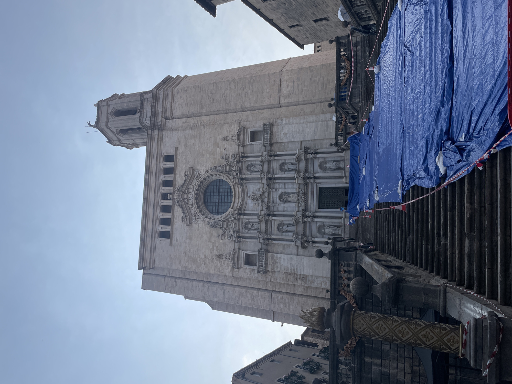 The image shows a low-angle view of the Girona Cathedral in Girona, Spain. A large blue tarp covers a significant portion of the wide stone staircase leading up to the cathedral's entrance. The tarp is secured with ropes and weighted objects, suggesting ongoing construction or preservation work. Red and white caution tape is visible along the sides of the stairs and around construction cones. The cathedral facade, made of light-colored stone, features intricate sculptures and a large rose window. A bell tower is visible to the right of the main facade. The sky is overcast. The surrounding buildings, made of stone, are also visible on either side of the cathedral, framing the grand staircase.