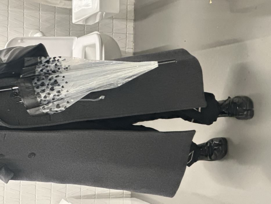 A single individual stands facing forward in what appears to be an indoor public restroom. The exact location in London, United Kingdom, cannot be determined from the image. The person is partially visible from the mid-torso downwards, wearing a dark grey, knee-length double-breasted overcoat and black trousers featuring a white angular design on the outer left leg. On their feet are black patent leather split-toe tabi shoes. The individual holds a closed transparent umbrella with scattered black heart patterns and a black handle. In the background, a white wall with a repeating 'M' or 'W' embossed pattern is visible. A white wall-mounted toilet with an open lid is positioned to the right of the person, and further right, a white wall-mounted sink is present. The floor is a uniform light grey. No explicit actions or interactions beyond standing are observable.
