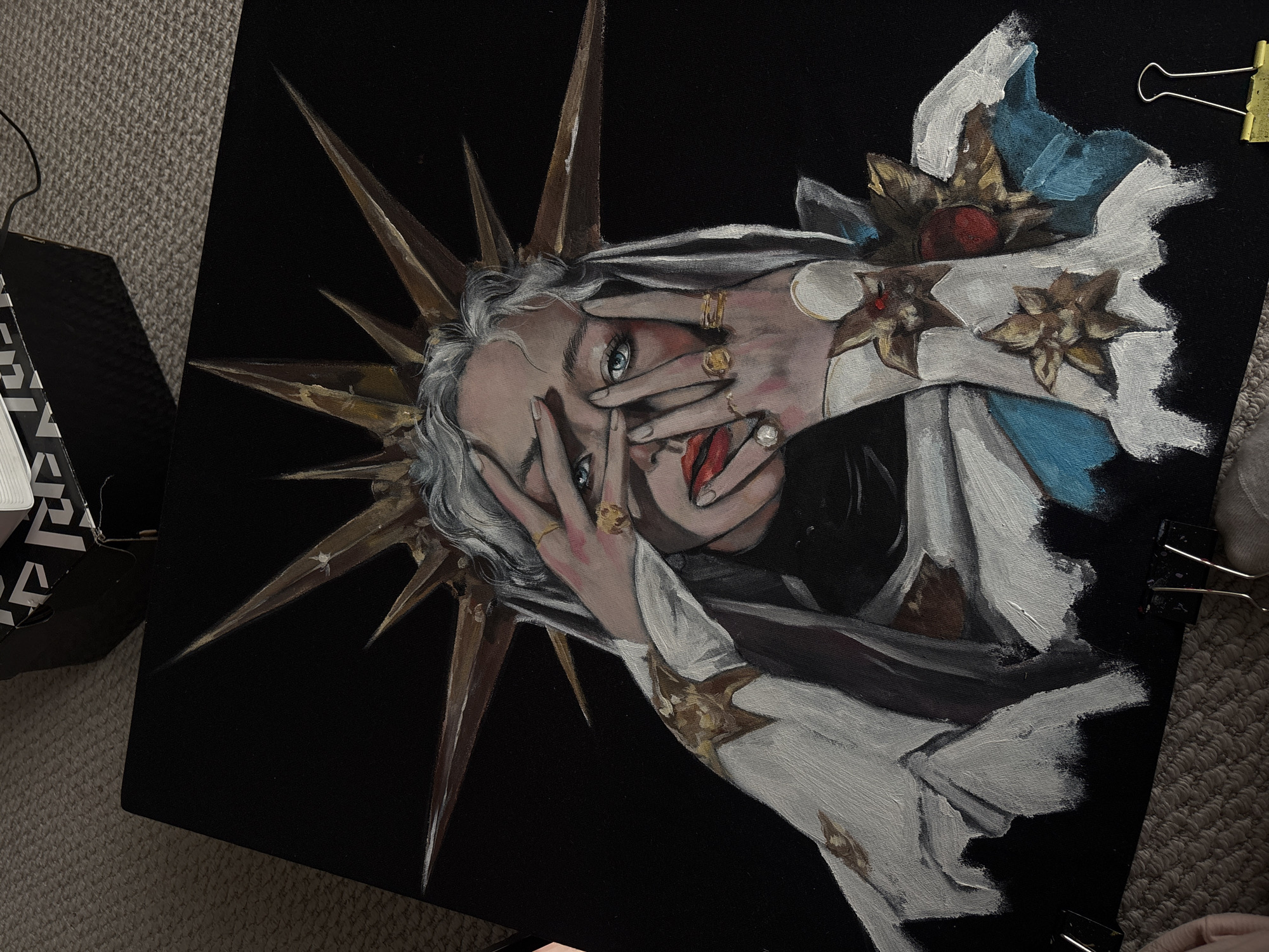 A close-up shot shows an oil painting on a black canvas. The subject is a pale-skinned person with white hair and blue eyes covering their face with their hands. Gold rings adorn several fingers. The person's red lips are visible. A golden crown with sharp, radiating spikes surrounds their head. The person is draped in white fabric with gold detailing and hints of blue near the top. The background is a solid black. The canvas is clipped on the right side with two silver binder clips and on the top right with another one, indicating it may be in progress. A portion of a black box with white text and geometric design is visible on the left side. The painting rests on a carpeted surface. The location is not Krasnoyarsk, Russia.