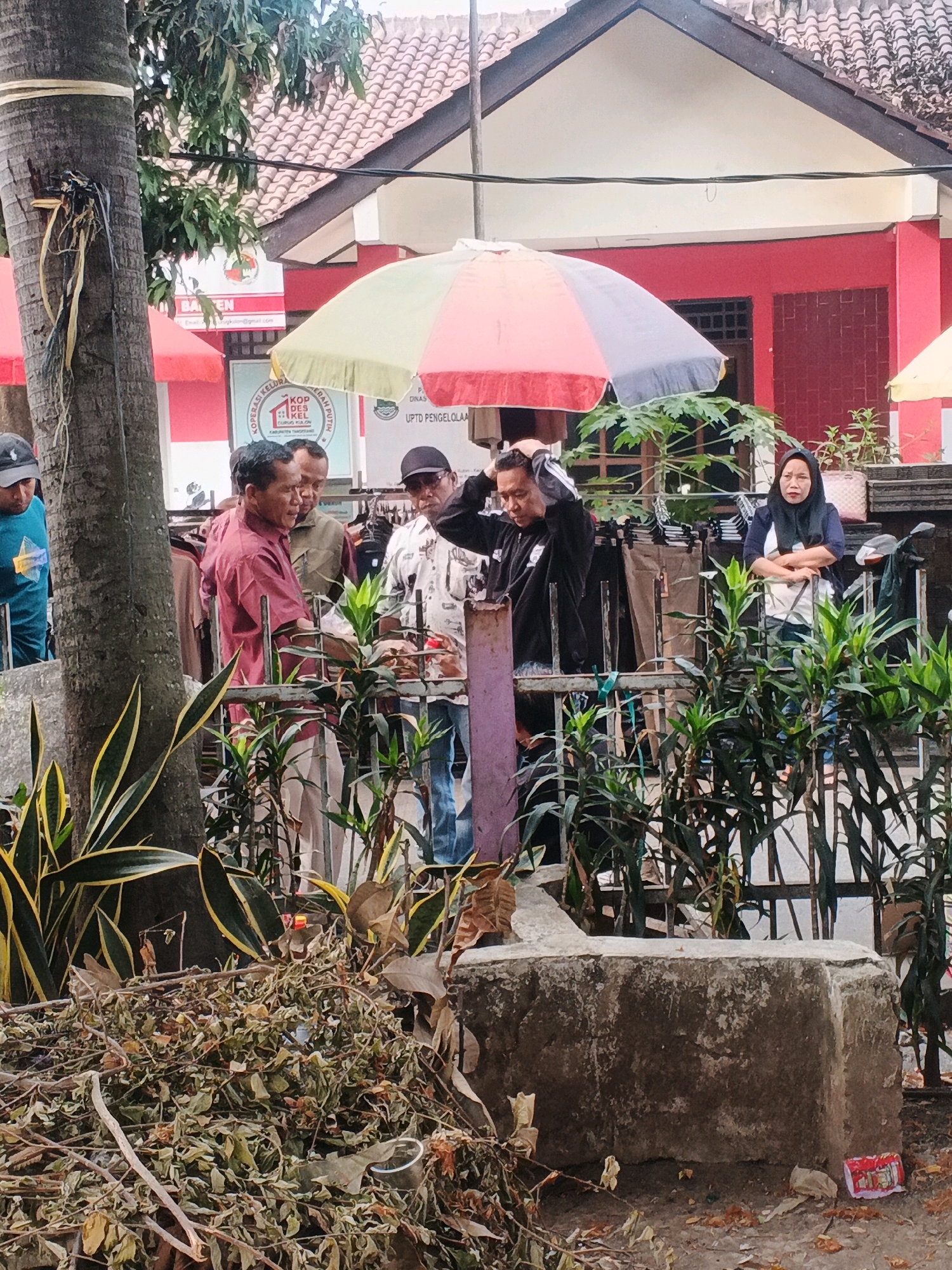 The image depicts an outdoor scene in Curug Kulon, Indonesia, likely near a market or government office. In the foreground, a pile of dried leaves and branches covers the ground, with a large concrete structure to its right. Behind this, a metal fence separates the foreground from a group of people.
There are at least five individuals visible. To the left, a person wearing a blue shirt and a dark cap looks down. Next to them, a man in a maroon collared shirt is speaking to another man wearing a light brown jacket. Further to the right, a man in a patterned short-sleeved shirt and glasses is standing, and a woman wearing a dark hijab and a white shirt is standing with her arms crossed. Behind the people, there are clothing racks with items hanging on them, suggesting a market stall or retail area.
Overhead, a colorful umbrella, predominantly yellow, red, and grey, is partially visible. In the background, a red building with a tiled roof is prominent. Signs are visible on the building and fence, including one with the text "BA EN" and another with "KOPERASI KEL". The environment appears to be a public area with trees and plants present.