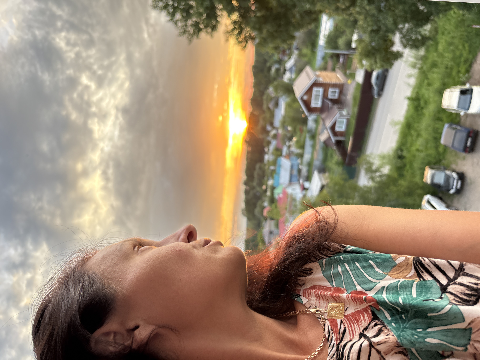 The media file shows a close-up, upward-angle shot of a woman's face and upper body, with a sunset visible in the background. The woman has dark hair and is wearing a necklace with a square pendant. She is looking upwards towards the sky, with a contemplative expression. Her clothing has a tropical pattern with large green leaves and some reddish-brown elements.
The background depicts a village or rural settlement at sunset. The sky is filled with dramatic clouds, tinged with the warm hues of the setting sun, ranging from bright yellow to orange and pink. Below the sky, there are houses with red roofs, some of them appearing to be wooden cottages, characteristic of a rural Russian setting. Trees and greenery surround the houses, and several cars are parked or driving on a road. The overall scene suggests a tranquil evening in a countryside location.
The time of day is clearly sunset, indicated by the warm colors in the sky and the low angle of the sun. The weather appears to be partly cloudy, with the sun breaking through the clouds. The location context provided is Chaikovsky, Russia, which aligns with the visual cues of the rural architecture and landscape. The woman's pose and gaze suggest a moment of reflection or appreciation of the natural beauty of the sunset. There is no visible text in the image.