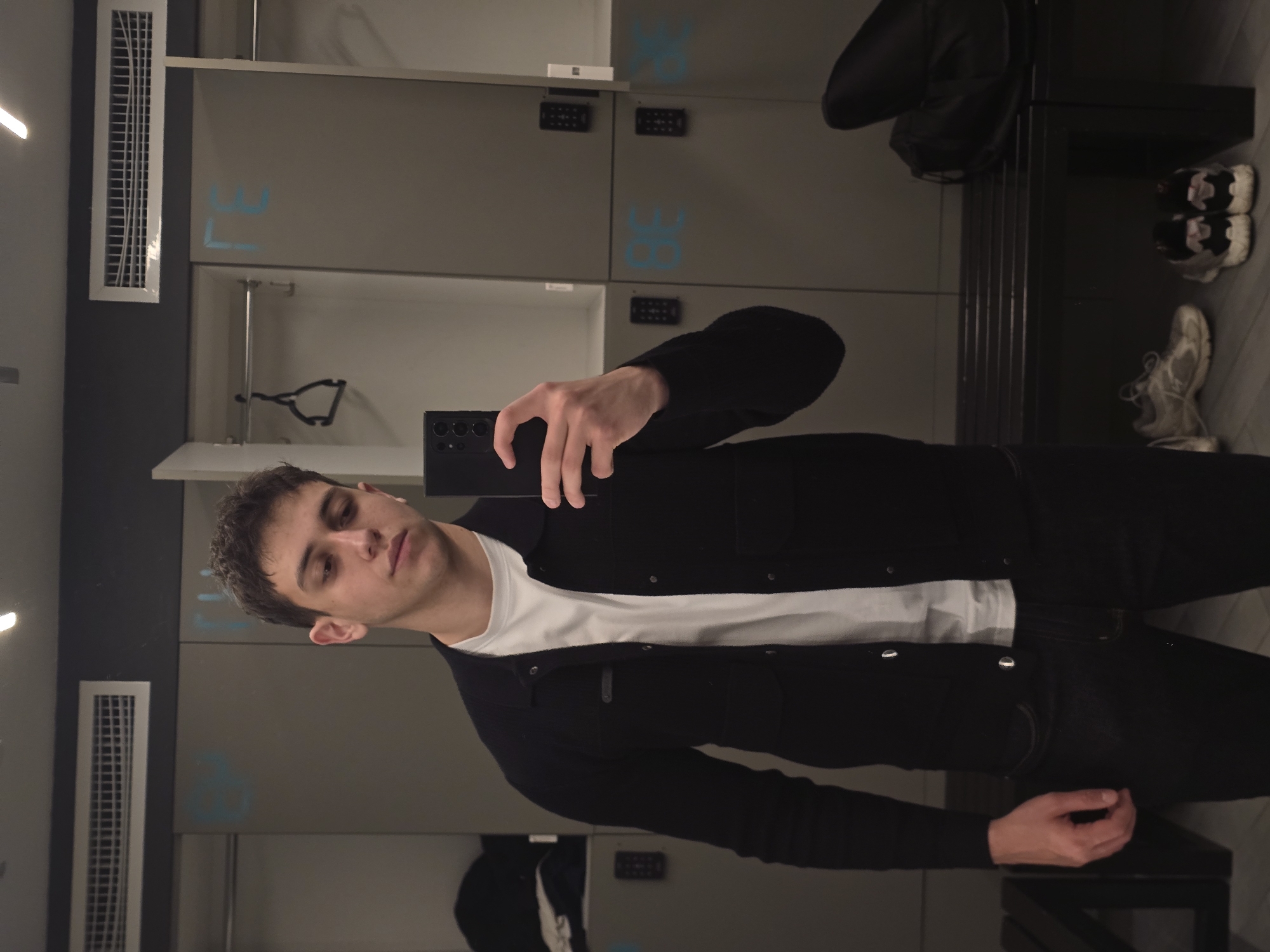 The image shows a person taking a selfie in what appears to be a locker room. The individual is wearing a white t-shirt, a black jacket, and black pants. The background includes lockers with numbers "37" and "38" visible. There are also a coat and a pair of shoes on a bench in the background. The location is likely a locker room, not specifically identifiable within the city of Yerevan, Armenia, based on the provided image. There are no visible interactions or specific actions occurring other than the person taking the selfie.