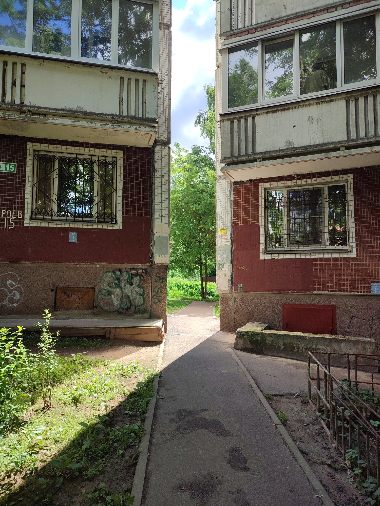 The image depicts the exterior of two adjacent apartment buildings in Vsevolozhsk, Russia, viewed from a pathway. The buildings appear to be constructed of concrete and tile, with some signs of wear and tear. Red tiles cover the lower portions of the buildings, while the upper sections are a lighter color, possibly painted concrete. Each building has several windows, some with metal grates. The pathway is asphalt, bordered by grassy areas and some overgrown vegetation. There is graffiti on the lower part of the left building. A metal railing is visible on the right side of the pathway, leading downwards. The sky above is partly cloudy with visible sunlight. The overall scene suggests a somewhat aged, urban residential environment.