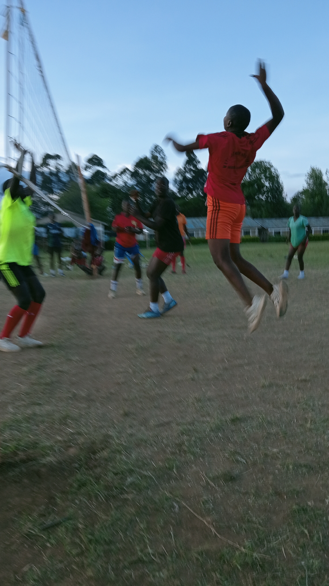 **Setting:** Outdoor volleyball game on a dirt and grass field in Ngenyilel ward, Kenya. The time of day appears to be late afternoon or early evening.
**People:** Several African individuals are present, engaged in or observing the volleyball game. One person is actively jumping to strike the ball. The clothing worn by the individuals varies, including sports attire in colors such as red, orange, yellow, green, and black.
**Objects:** A volleyball net is visible on the left side of the frame. The background shows vegetation and buildings in the distance.
**Actions:** The primary action is a person in mid-air, reaching up to hit a volleyball. Other individuals appear to be in various states of readiness or anticipation related to the game.
**Environment:** The surrounding environment consists of grass, dirt, trees, and some buildings that are likely residential or school buildings in the background. The lighting suggests the photograph was taken during dusk.
