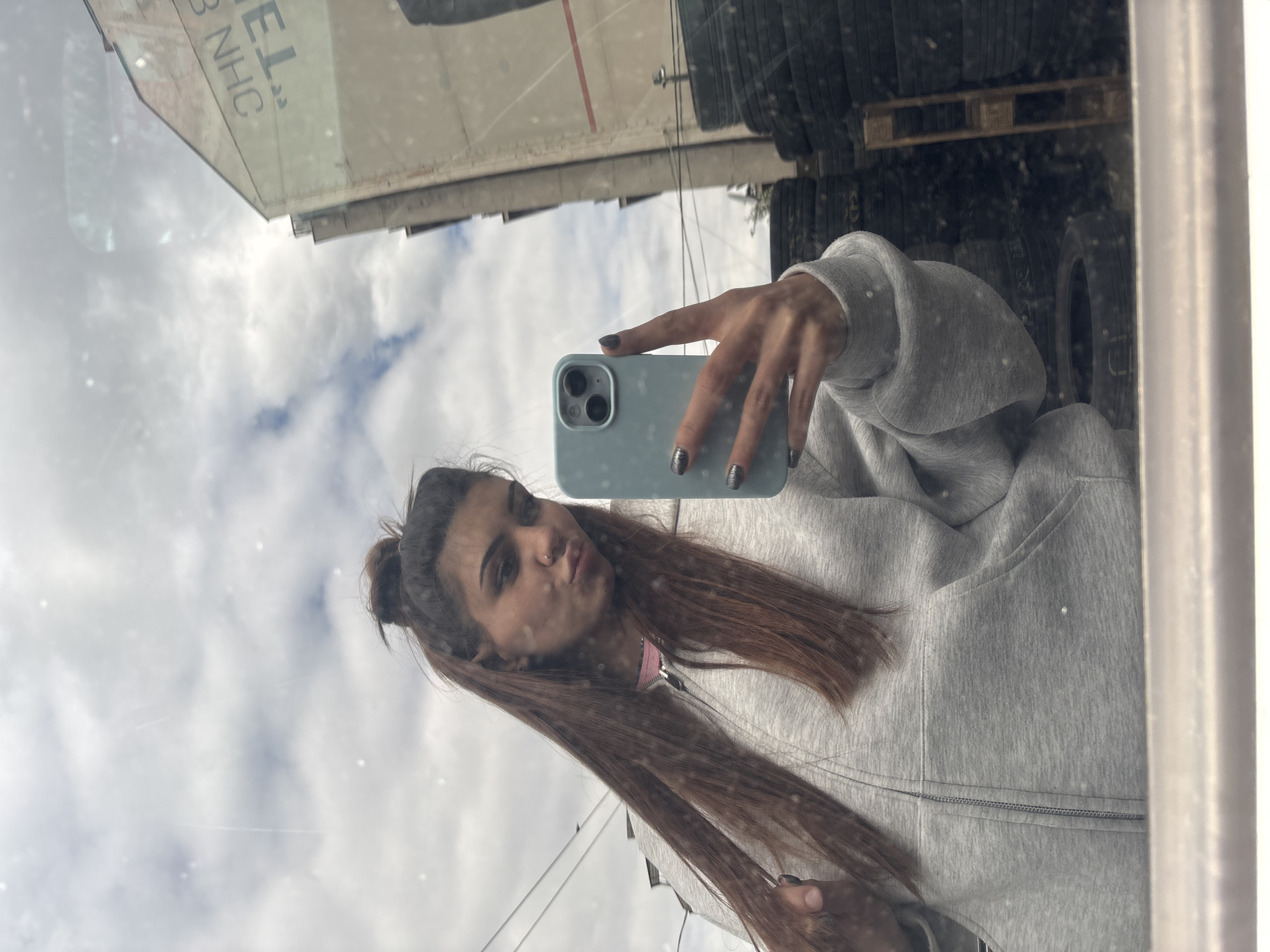A young woman takes a selfie in a reflective surface, possibly a window, in what appears to be an industrial area of Sofia, Bulgaria. The reflection shows stacks of tires and the side of a building with the letters "EL" and "NHC" visible. The sky reflected in the window is cloudy. The woman is wearing a gray hooded sweatshirt and has her hair styled in a partial updo. She is holding a blue smartphone and puckering her lips for the selfie. Her nails are painted a dark, metallic color.