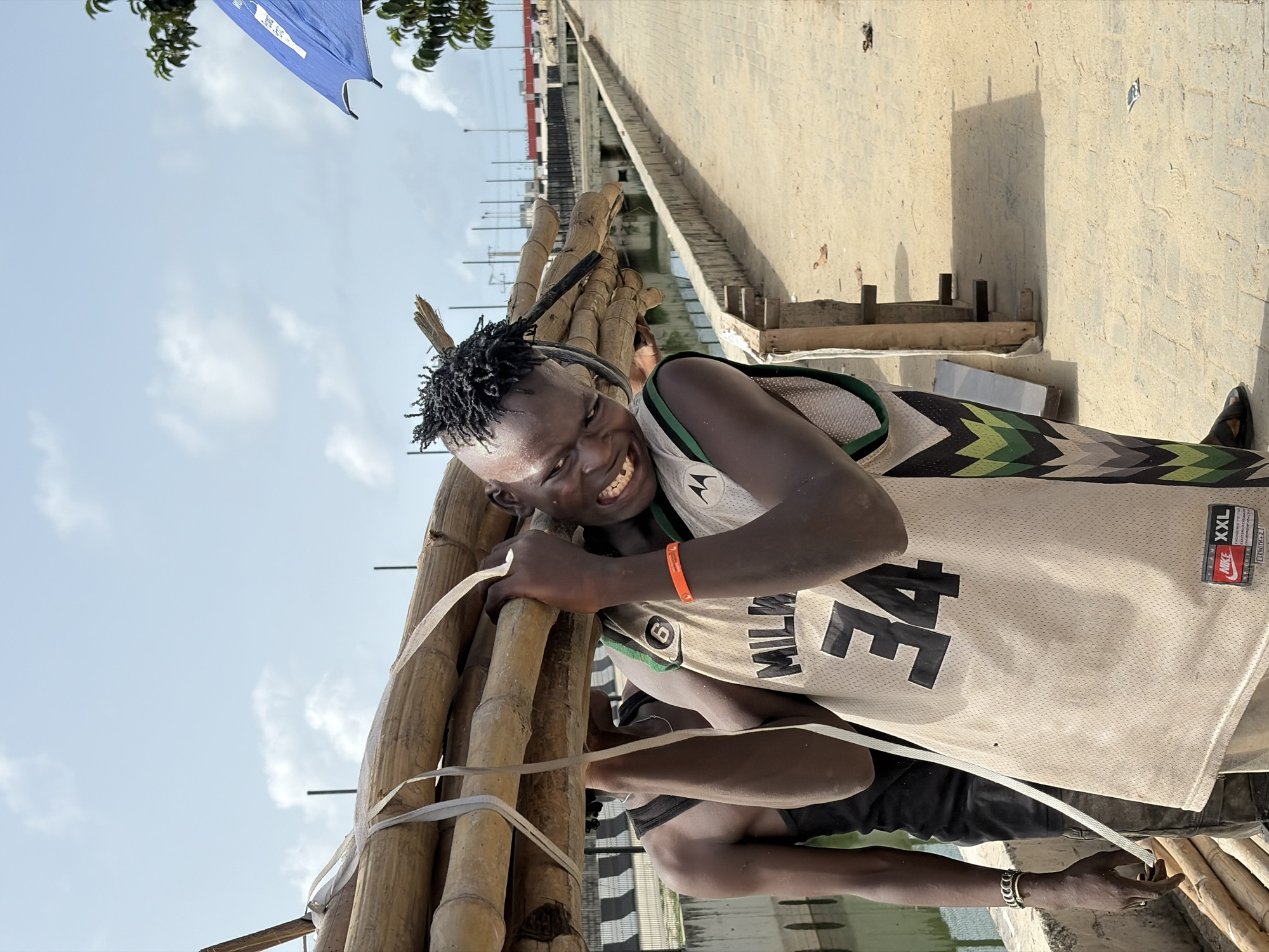 A man stands in Igboefon, Nigeria, carrying a bundle of bamboo. He wears a Milwaukee #34 basketball jersey, black shorts, and sandals. An orange bracelet adorns his right wrist. The bamboo is secured with white plastic strapping. A bridge, water, and a blue banner are visible in the background. The picture is taken in daylight.