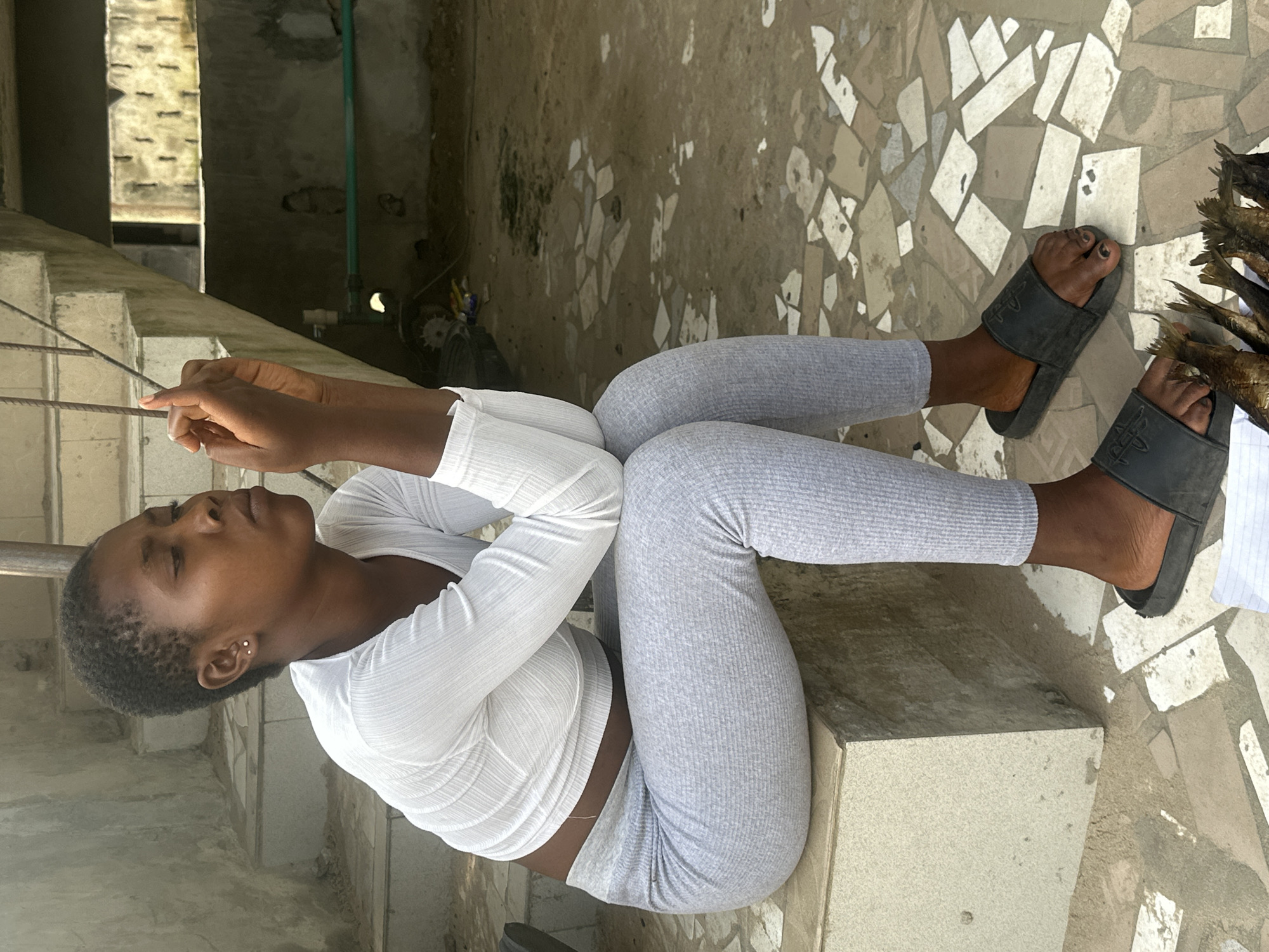 A young woman with short hair sits on a concrete ledge in Badagry, Nigeria. She is wearing a white long-sleeved crop top and light gray ribbed leggings, with black slides on her feet. Her arms are crossed in front of her, and her head is tilted down as she looks at her hands, which are clasped in front of her face. In front of her, on the tiled ground, is a pile of dried fish. The background shows a concrete staircase and a wall with exposed pipes and wiring. The scene appears to be an outdoor, possibly semi-constructed, area.