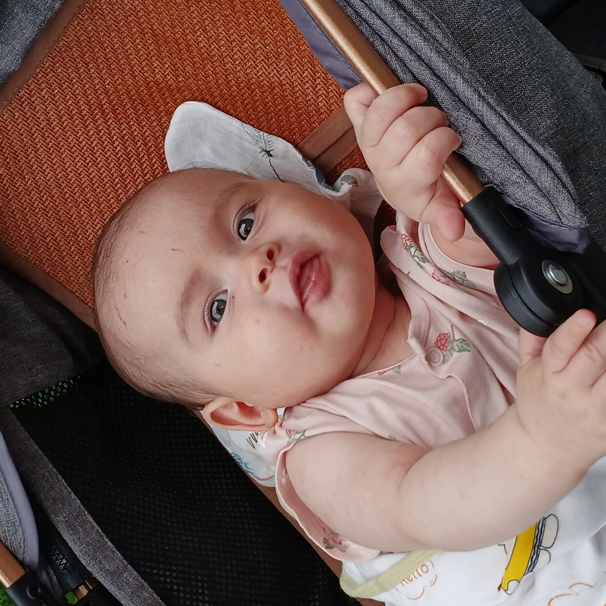 A baby lies in a stroller. The baby is fair-skinned with dark eyes and is wearing a pink top with a white and yellow undershirt. The baby is holding onto the stroller's handle with both hands and has their tongue slightly protruding. The stroller has a brown woven seat, a grey canopy, and black mesh underneath. The location is Shunde District, China. There is no other information that can be determined about the environment.