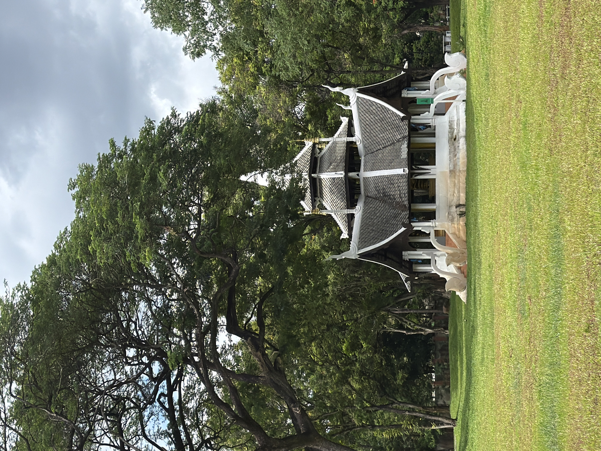 In Chiang Mai City Municipality, Thailand, an open grassy area is dominated by a traditional Thai pavilion with a multi-tiered, dark grey roof and white decorative trim. The pavilion is situated amidst lush green trees, with a large, sprawling tree with dark branches in the foreground on the left. The sky above is overcast with grey clouds. The pavilion has a white base adorned with white ornamental carvings resembling stylized birds or mythical creatures on either side of the entrance stairs. The surrounding environment is a park-like setting with well-maintained green grass. No people are visible in the scene.