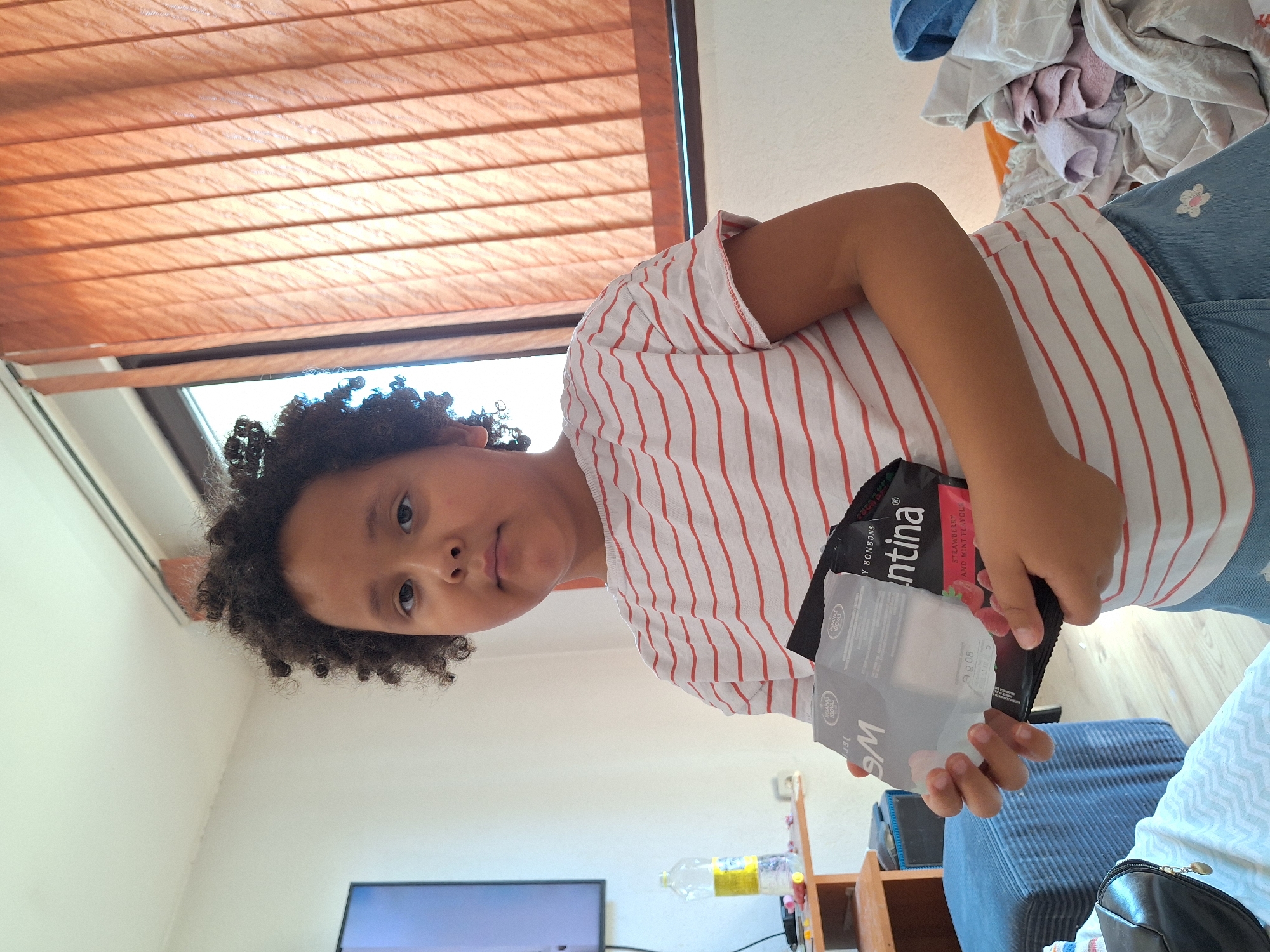 A young girl with dark, curly hair and brown eyes is standing in a room, holding two packages. She is wearing a white t-shirt with thin red stripes and a blue denim skirt with a small floral pattern. The packages she is holding are a small white box and a black bag with text that appears to be in a foreign language, possibly including the word "Valentina" and "Strawberry and Mint Flavour."
The room appears to be a domestic setting. To the left of the girl, a television screen is visible. Further left, a shelf holds a plastic bottle of water with a yellow label, and other miscellaneous items. In the background, a window is covered by orange vertical blinds, with bright light coming through. To the right of the girl, a pile of folded laundry, including towels and blankets in various colors, is visible. A blue upholstered stool is in the lower left corner of the image. The flooring is a light-colored wood. The overall impression is a casual, indoor scene, likely in a home or dormitory, in Blagoevgrad, Bulgaria.