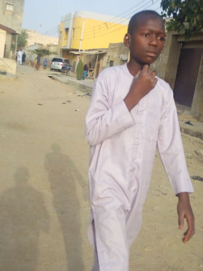 An outdoor scene in Garun Gawa, Nigeria, depicts a young male in the foreground walking on an unpaved, light brown dirt road. The individual, estimated to be 10-14 years old, wears a light purple or off-white long-sleeved tunic. His right hand is raised to his chin/neck area. His gaze is directed slightly to his right.
In the mid-ground, a silver SUV and a motorcycle are parked on the left side of the road. Several other individuals are visible in the distance: a group of adults in light-colored traditional attire, a woman in a dark green garment near a makeshift stall or structure, and a child near her. Two goats are also present on the dirt road.
The background features a mix of architecture. On the left, a light-colored building with a visible window is partially seen. Towards the center-right, a prominent two-story building with a bright yellow facade and a curved silver architectural element stands out. Other dilapidated structures with wooden frames and dark openings are scattered throughout the background. Utility lines are visible against the clear, light blue sky. Two distinct human shadows are cast on the dirt road to the left of the primary subject, indicating a high sun position.
