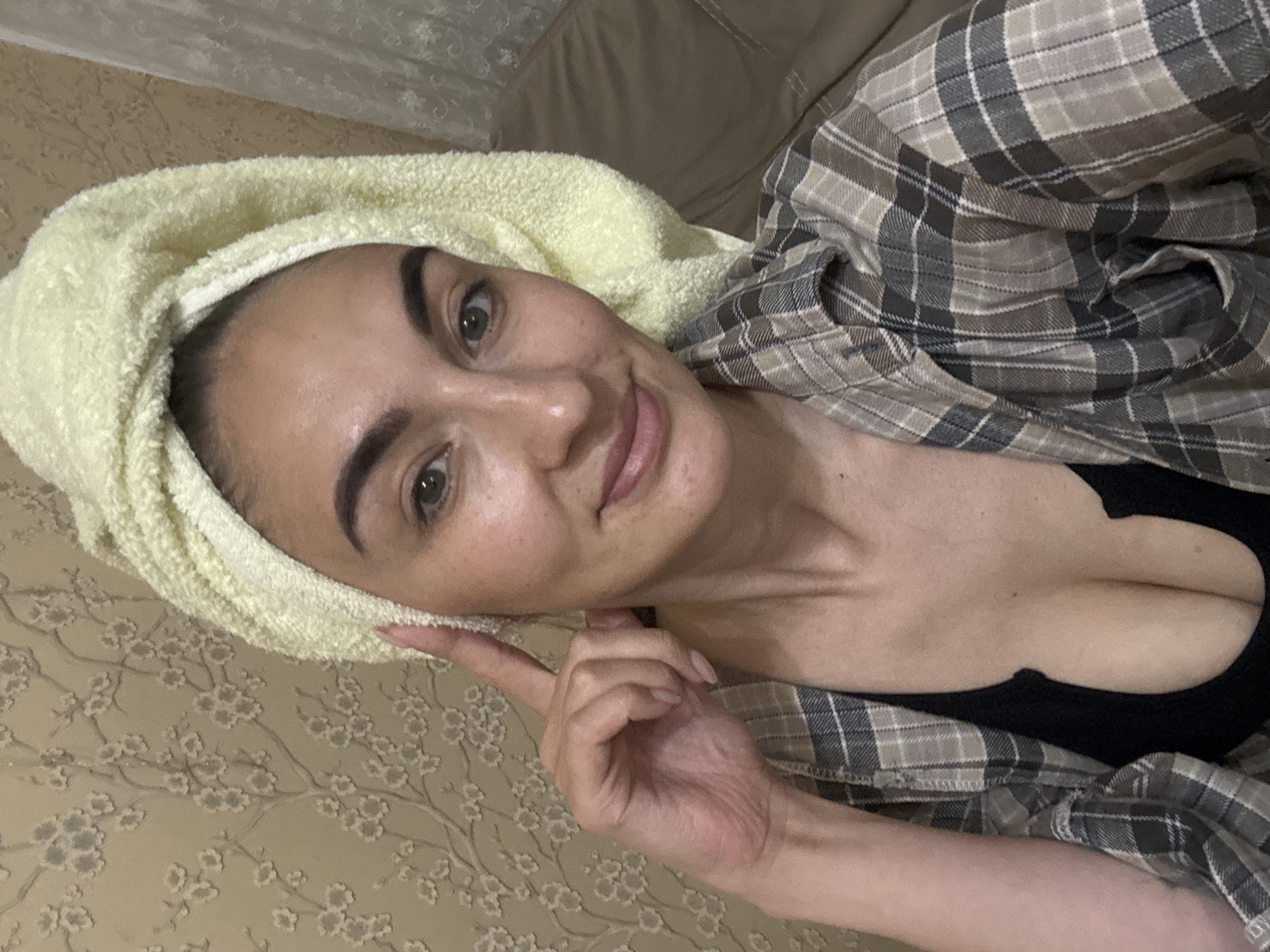 A woman is reclining indoors, wearing a black top and a plaid flannel shirt. Her hair is wrapped in a yellow towel. She is pointing at the towel with her right hand. The background consists of a bed with a patterned bedspread and a tan headboard. The location in the city of Sochi, Russia is not discernible from the visual information.