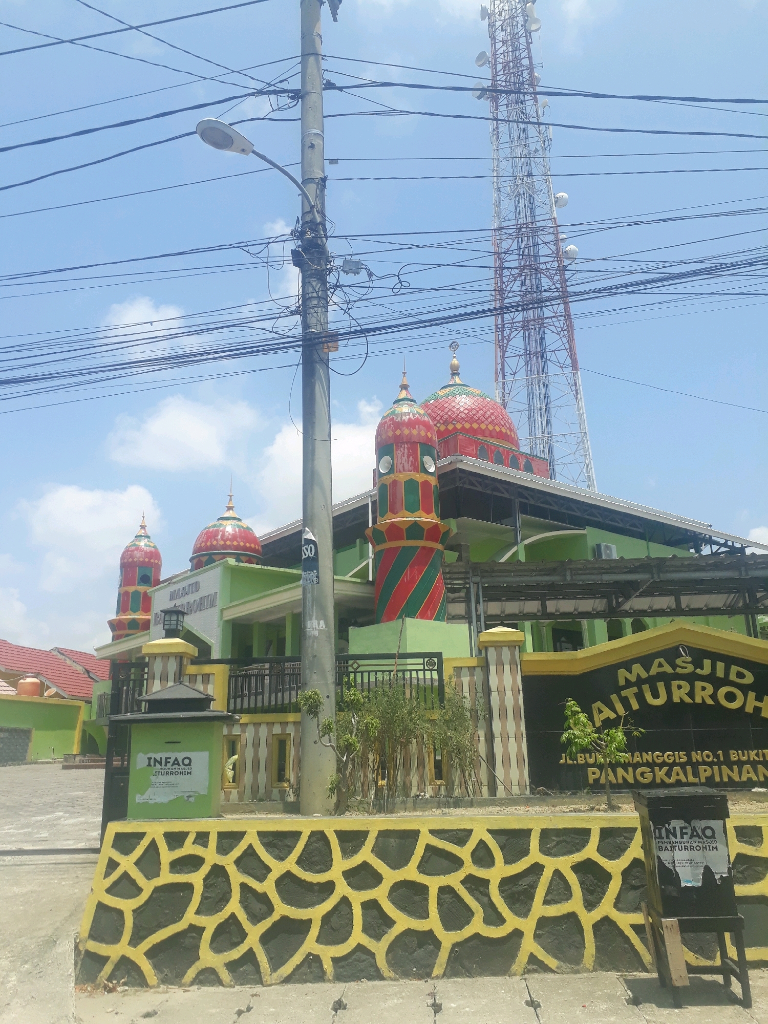 The image shows the Masjid Baiturrohim mosque located at Jl. Bukit Manggis No.1, Bukit Manggis, Pangkalpinang, Indonesia. The mosque is painted in green with red and green striped minarets topped with golden finials. Two prominent red domes are visible on the main building. In front of the mosque, there is a decorative wall with a black and yellow abstract pattern. A street lamp on a utility pole with many wires is in the foreground, partially obscuring the view of the mosque. Behind the mosque, a tall telecommunications tower is visible against a bright blue sky with scattered clouds. A sign with the mosque's name and address is mounted on a yellow structure. There is also an "INFAQ" donation box. The overall scene depicts a sunny day in an urban or suburban area, with the mosque being the central focus.