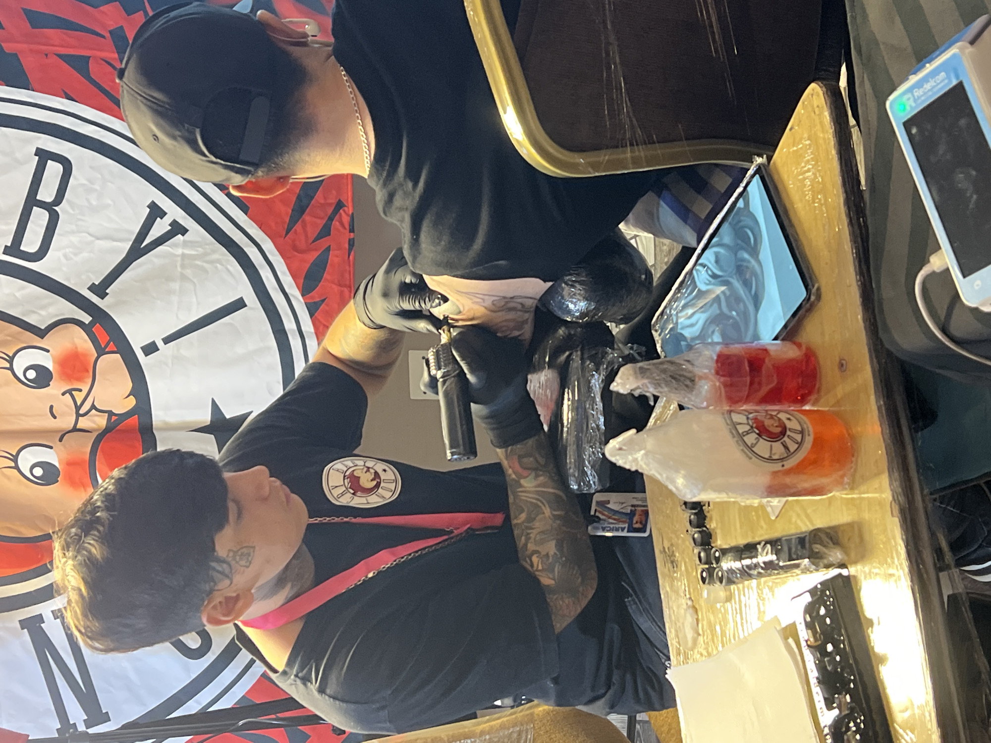The image depicts a tattoo artist, identified by a tattoo on their face, in the process of inking a client's arm. The artist is wearing black gloves and a black t-shirt with a circular logo. The client, wearing a black baseball cap and a silver chain, is seated and receiving the tattoo on their upper arm. A banner with a cartoon character and black and red designs serves as a backdrop. On a table in the foreground, there are bottles of tattoo ink, a roll of paper towels, a tattoo machine, and a tablet displaying a design. The presence of a lanyard with a name tag, partially visible in the lower part of the frame, suggests a public event or convention. The distinctive graphic style of the banner, particularly the caricature of a baby with rosy cheeks and large eyes, is a recurring motif associated with the "Baby Rock" tattoo studio, which has a presence in Arica, Chile. The lighting suggests an indoor setting with artificial light sources.