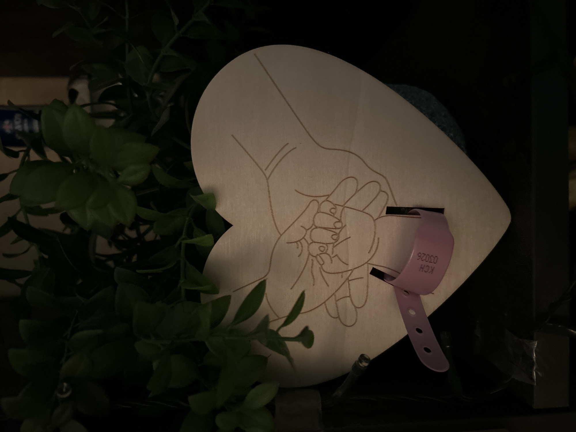The image shows a wooden heart-shaped ornament with an engraved outline of hands holding a baby's hand. A pink wristband with the text "KCH 03026" is attached to the ornament. The ornament is positioned in front of some artificial green foliage, and it is likely a keepsake. The scene is dimly lit, suggesting it might be indoors in a home or a decorative display. The location in Sofia, Bulgaria, is not depicted in the image.