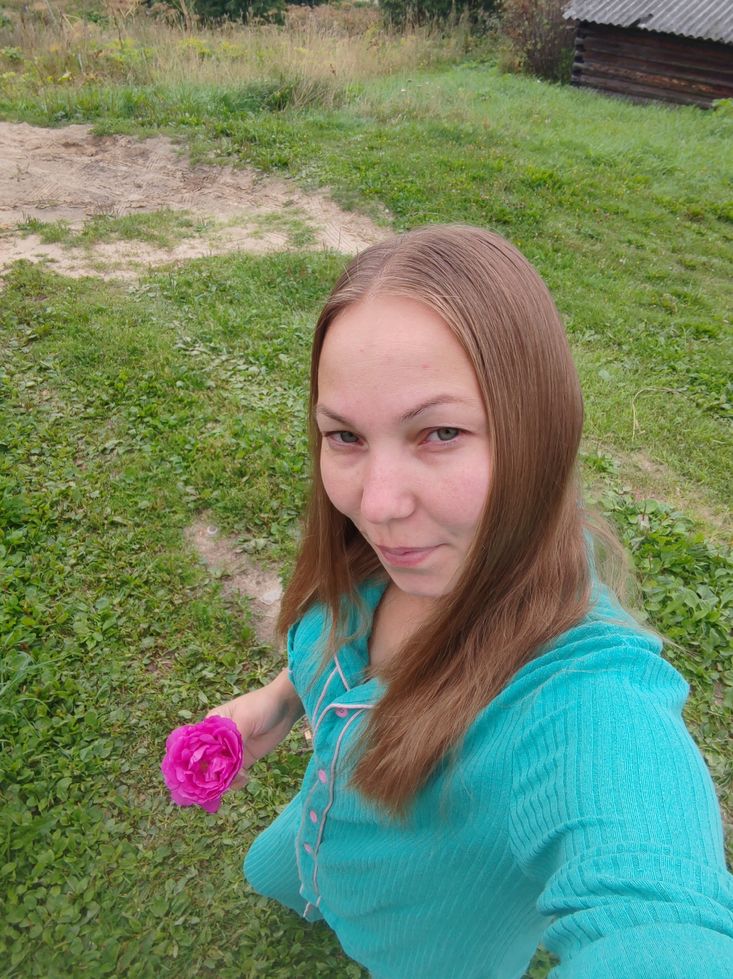 A young woman with long, light brown hair is holding a pink rose in her left hand. She is wearing a turquoise, ribbed garment with pink trim and buttons. She is looking towards the camera with a slight smile. The background is a grassy area with a dirt path and a rustic wooden building with a corrugated roof. The scene appears to be outdoors in a rural setting, possibly in the city of Chuchlem, Russia, given the architectural style and vegetation.