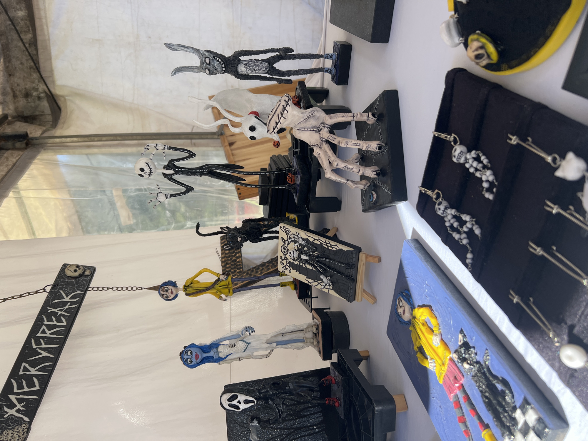 The image displays a selection of handcrafted figures and jewelry. The figures appear to be inspired by various films, including “The Nightmare Before Christmas”, “Coraline” and “Corpse Bride”. The figures are displayed on a white surface in an indoor setting. The jewelry includes skeleton-themed earrings and other pieces arranged on black display pads. The objects suggest the scene is a craft fair or market stall, specializing in artistic and potentially handmade goods. The “MERRYFREAK” sign is also on the counter, and the jewelry displays “Jack Skellington” and “Zero” from The Nightmare Before Christmas. The exact location within Buenos Aires, Argentina, cannot be determined from the visual data provided.