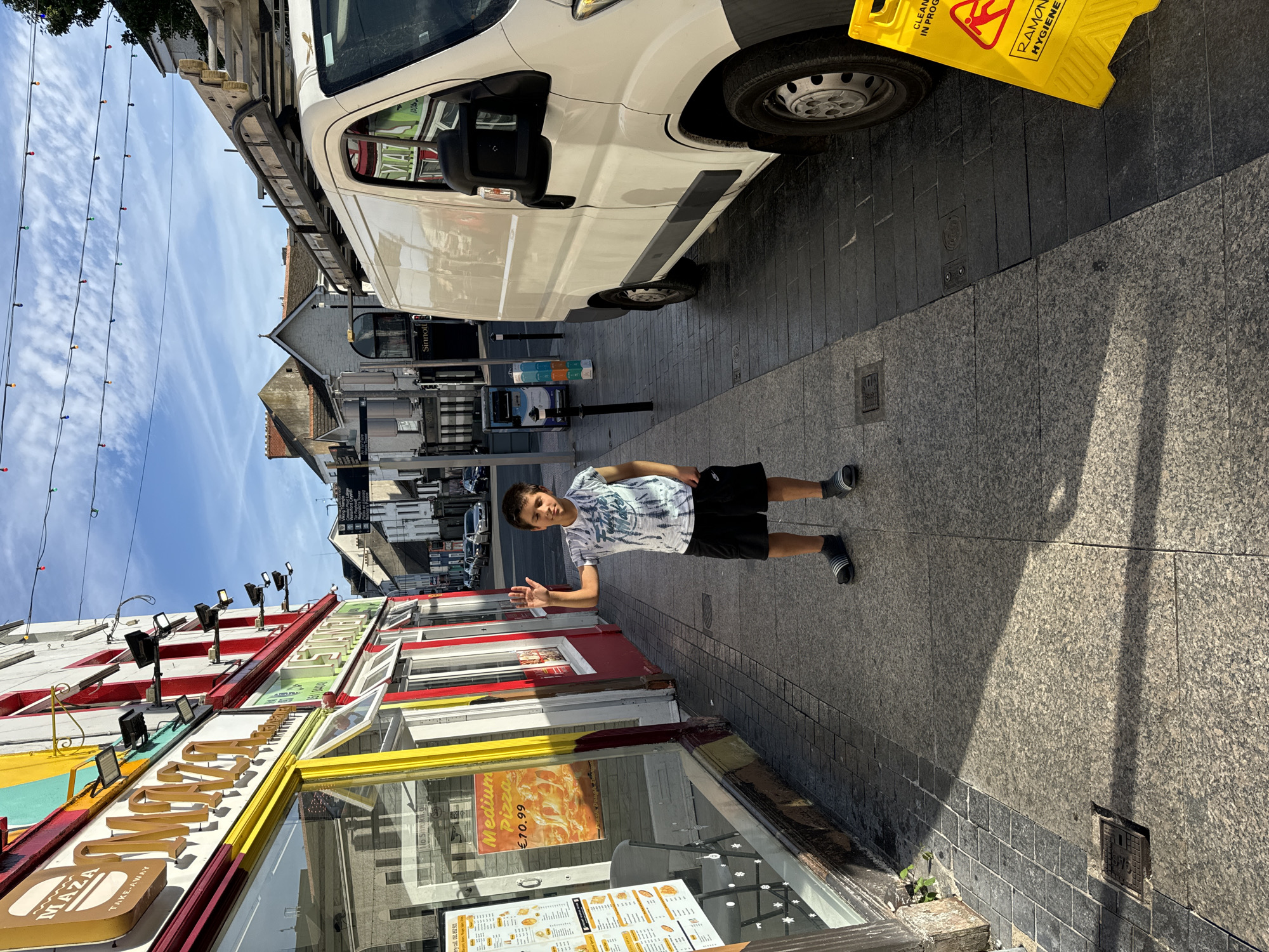 A young boy, approximately 10 years old, stands on a sidewalk in front of a storefront. He is wearing a tie-dye t-shirt and black shorts, and he is waving with his right hand. The storefront is colorful, with a large "MAZA TAKE-AWAY" sign. A white van with a ladder on top is parked on the street beside the sidewalk. A yellow caution sign indicating "CLEANING IN PROGRESS" is visible on the ground near the van. The scene appears to be in a commercial area, with other buildings and streetlights visible in the background. The sky is blue with some clouds, and sunlight is casting shadows on the sidewalk. Based on the signage and the overall context, this scene is likely located in Waterford City, Ireland.