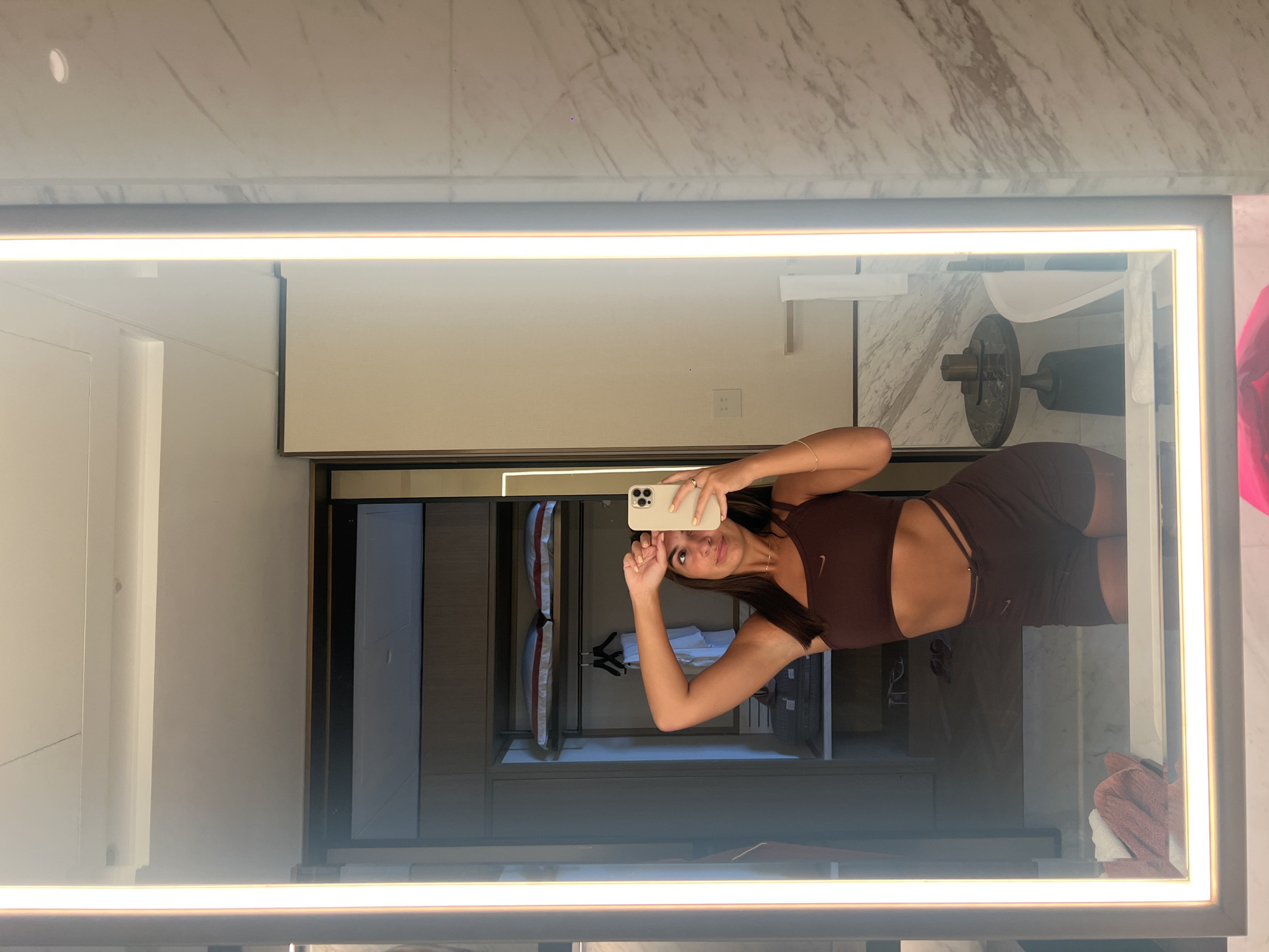 A woman is taking a selfie in a bathroom mirror. She is wearing a brown Nike sports bra and matching shorts. The bathroom has marble walls, a white sink, and a dark wood closet. The setting suggests a high-end hotel or residence in Dubai, United Arab Emirates.