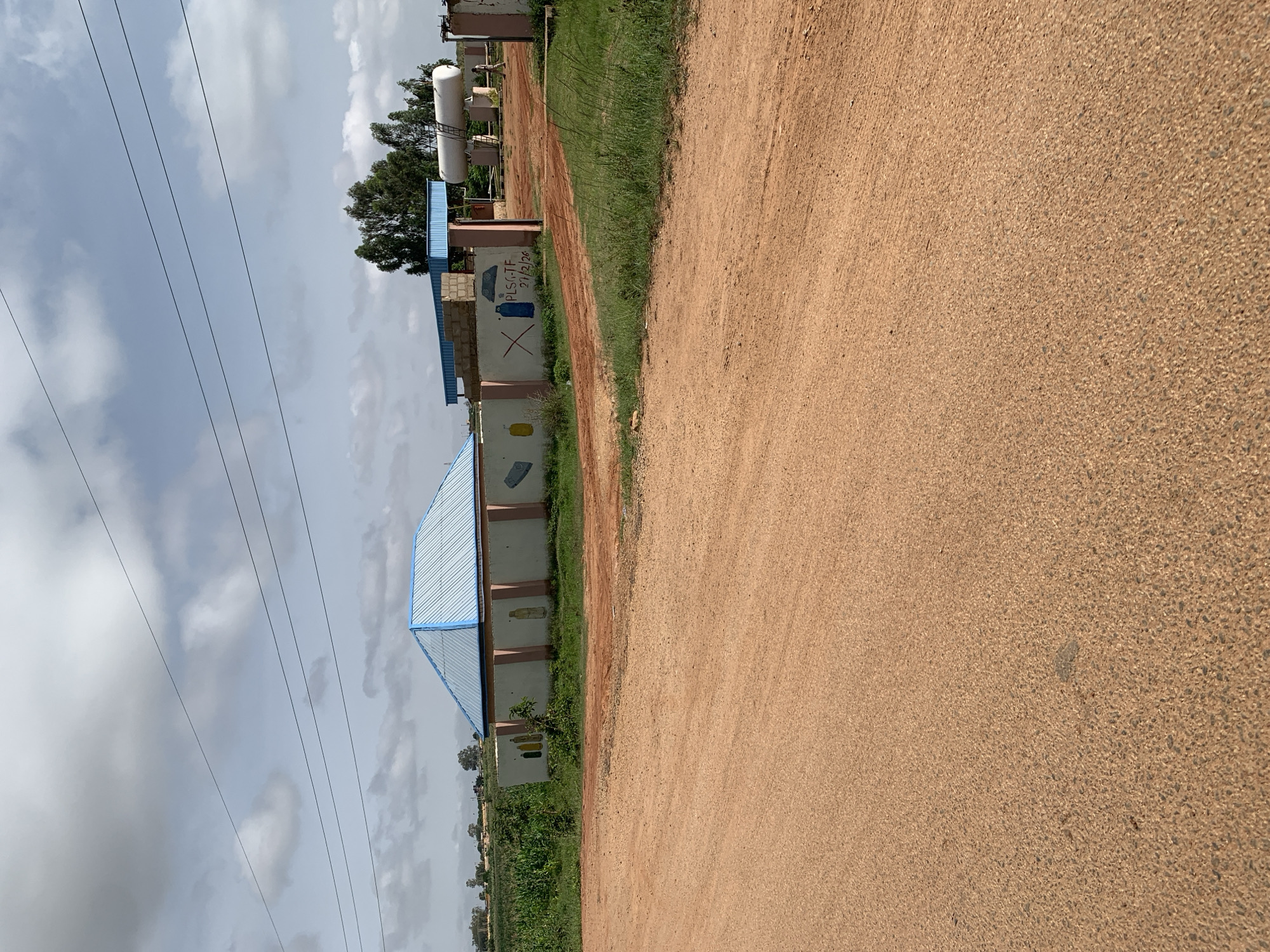 The image depicts an outdoor scene in Nabatum, Nigeria. In the foreground, a dirt road takes up a significant portion of the frame, with patches of green grass growing along its edges. Behind the road, a low, light-colored wall runs horizontally across the mid-ground. The wall is adorned with simple, colorful painted images, including what appear to be bottles and a gas cylinder. A blue and white building with a peaked roof is visible behind the wall on the left side of the image. To the right of this building, a more open area reveals a large white cylindrical tank, likely a storage tank, elevated on supports. A person is seen walking near this tank. In the background, lush green vegetation and trees are present, under a partly cloudy sky. Power lines are strung across the upper portion of the image.