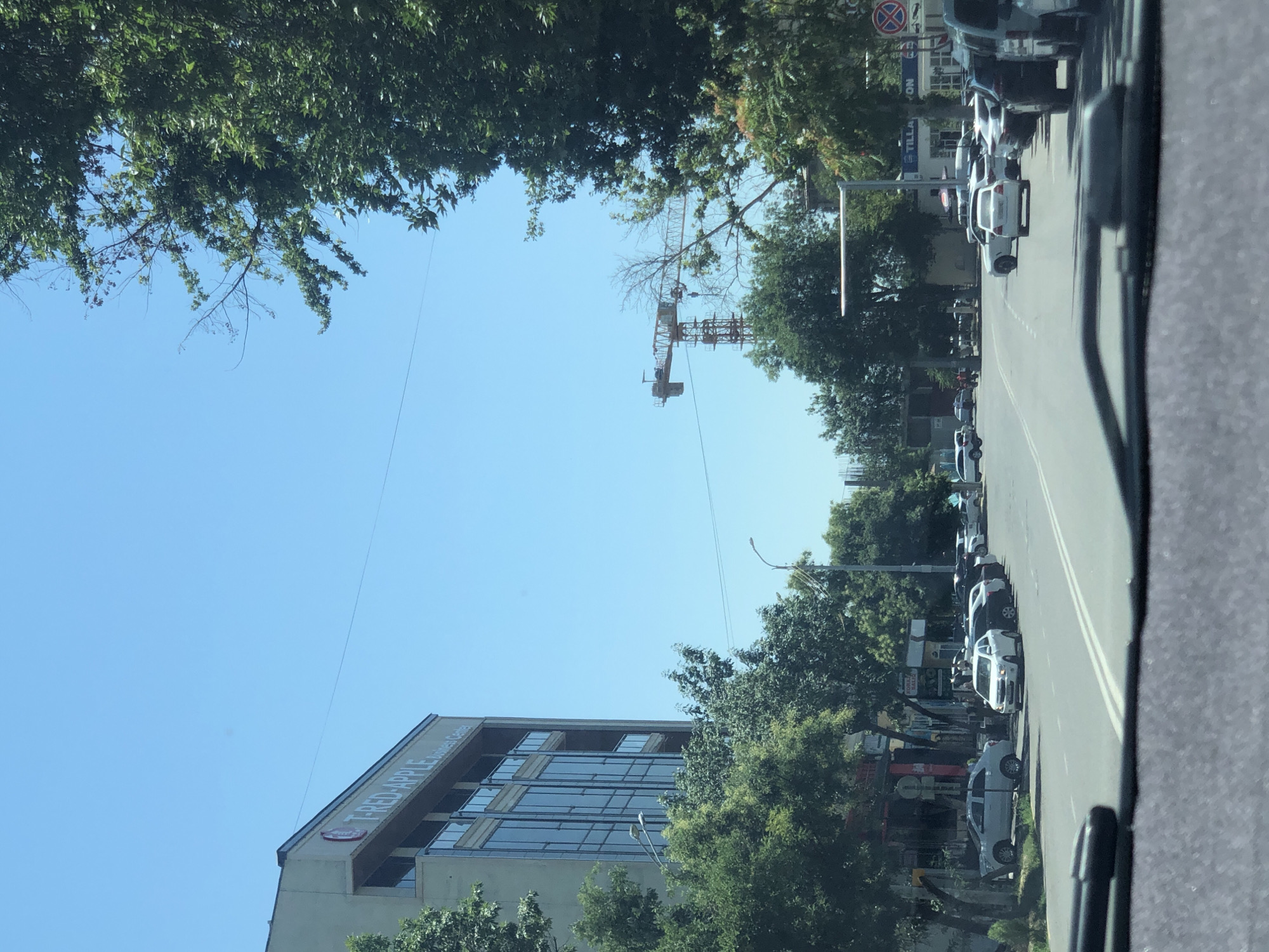 The image depicts a street scene in Katta Chilanzar-2 MFY, Uzbekistan, on a clear, sunny day. A multi-story building with a sign reading "T-RED-APPLE Business Center" is visible on the left. The building has a modern facade with large glass windows. Trees line the street, some obscuring parts of the buildings. A construction crane is visible in the distance. Several cars are parked along the side of the road and on the street. The road has lane markings. The viewpoint appears to be from inside a vehicle looking forward through the windshield. The overall environment suggests a commercial or mixed-use urban area.