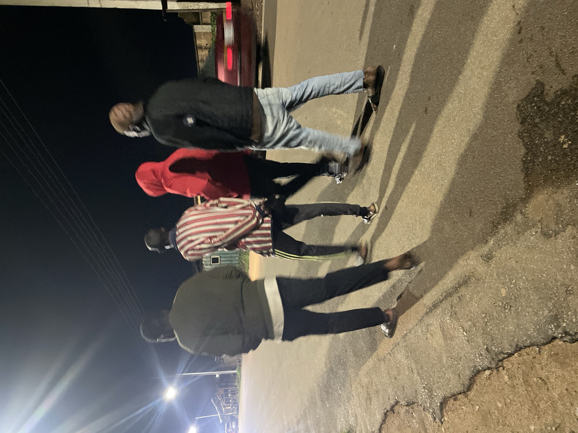 Four individuals are walking away from the camera on a paved road at night in Barakin Lerawi, Nigeria. The road appears to be an unlit street, possibly a residential or less developed area, with a cracked and uneven surface. Streetlights are visible in the background, casting long shadows of the people and creating a dimly lit atmosphere.
The person closest to the camera on the left is wearing a dark long-sleeved shirt and black pants, with sandals on their feet. To their right, another person is wearing a red hooded jacket and dark pants, also with sandals. Next to them, a person in a vertically striped shirt (red and white) and dark pants with yellow stripes is walking. The furthest person on the right is wearing a black jacket, light blue jeans, and sandals. The group appears to be casually strolling. In the background, a red car with illuminated taillights is partially visible, suggesting traffic or parked vehicles. Buildings and what might be wooden structures are present further in the background, partially obscured by darkness and distance. The overall scene depicts a nighttime street environment in a Nigerian locality.