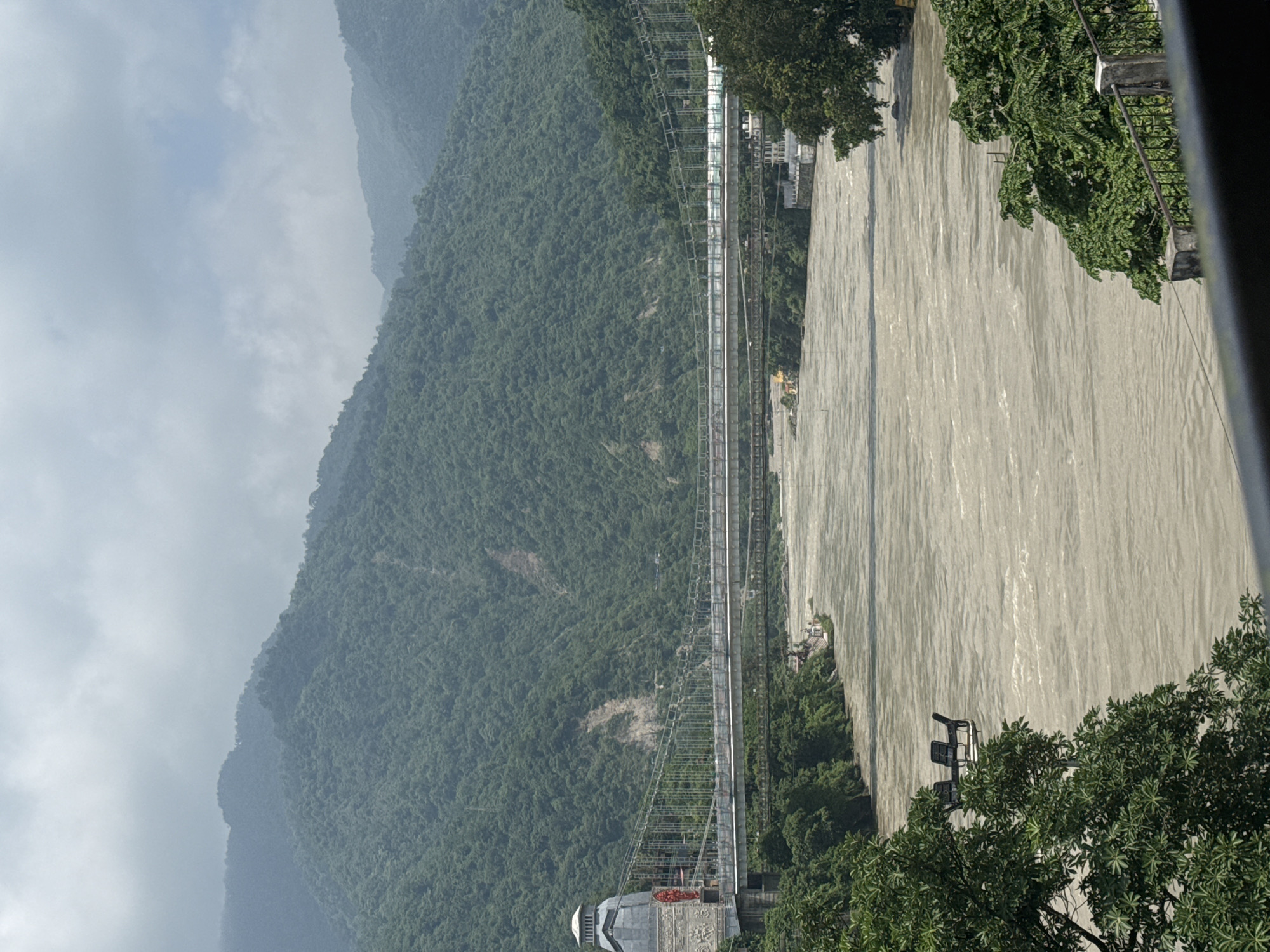 The image captures a wide, muddy river flowing through a valley, flanked by lush, green mountains. A suspension bridge, the Lakshman Jhula, spans the river. On the left bank, a decorative tower with intricate carvings stands at the entrance to the bridge. In the foreground, lush green trees frame the scene, and to the left of the river, a few empty chairs are visible on a platform. The sky is overcast, with clouds obscuring the mountaintops. The overall scene depicts a natural landscape with significant man-made structures.