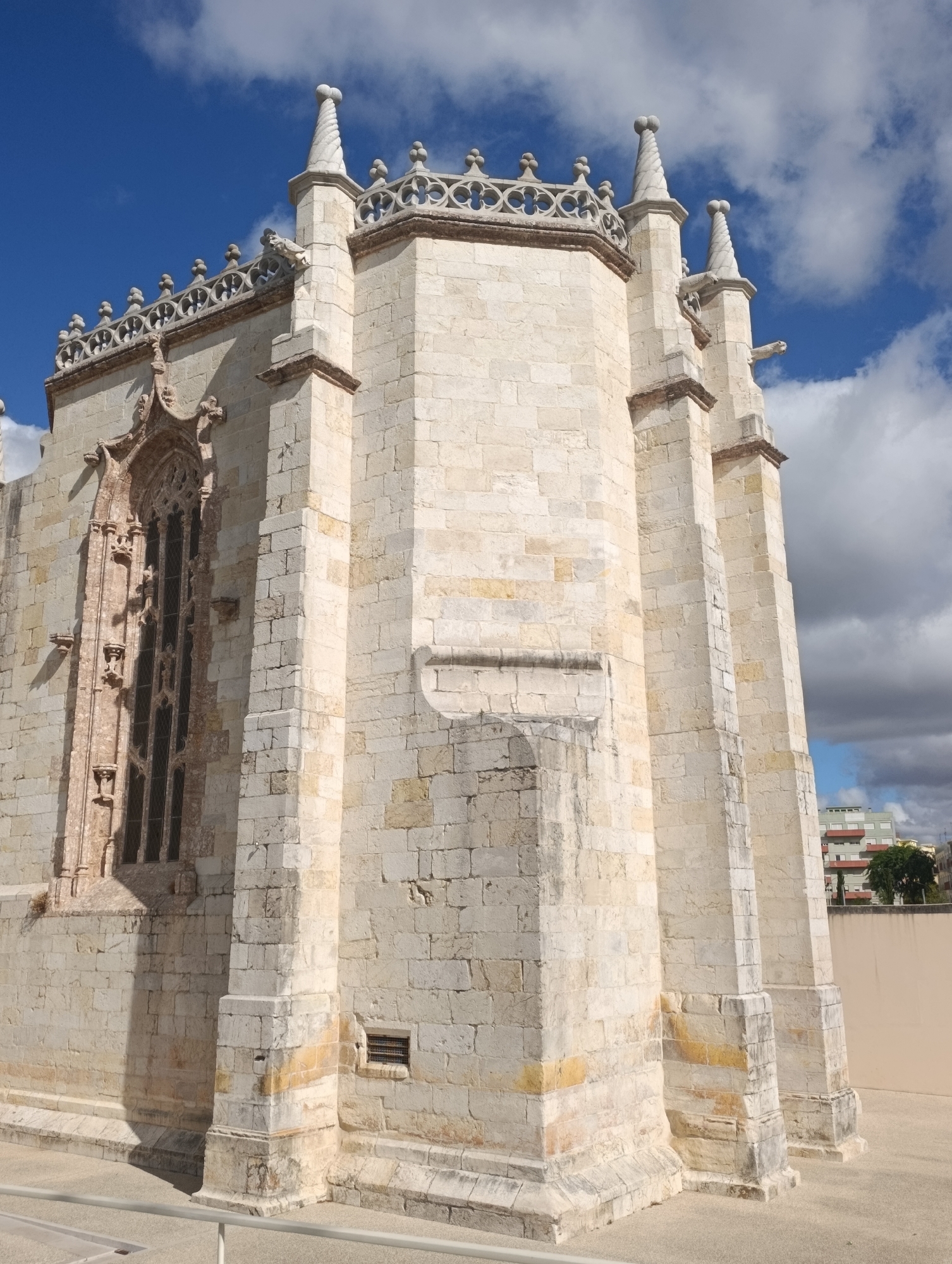 This image shows the Jerónimos Monastery in Belém, Lisbon, Portugal. The monastery is a UNESCO World Heritage site and a prime example of Manueline architecture. The image is taken from a low angle, looking up at the intricate stonework of the monastery. The building is made of light-colored stone and features Gothic and Renaissance architectural styles. The sky is blue with some clouds, and the sun is casting shadows on the building. In the background, there is a glimpse of a modern building and trees. The image is a high-quality photograph, likely taken with a smartphone or digital camera.