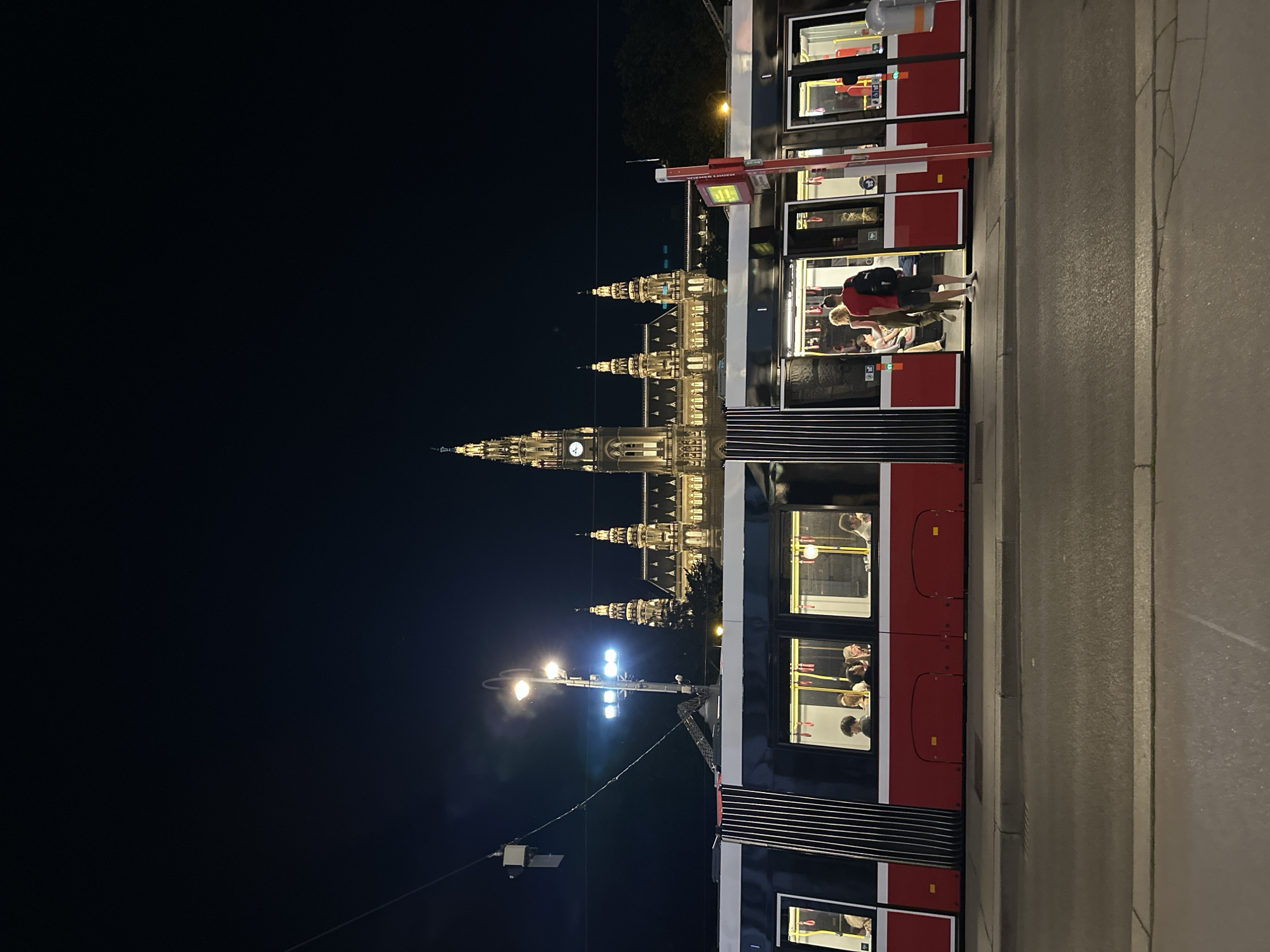 The image shows a night scene in Vienna, Austria, with the Rathaus (City Hall) illuminated in the background. A red and black tram with several passengers inside is stopped at a tram stop. Two people are standing at the open doors of the tram, one appearing to be boarding and the other waiting. The street in front of the tram is paved, and a sidewalk runs alongside it. The sky is dark and clear, indicating nighttime.