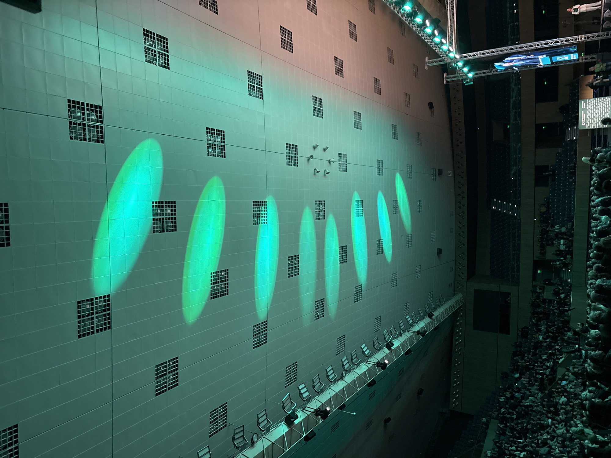 The image depicts a large indoor event within the Palace of Culture and Sports in Varna, Bulgaria. The scene shows a spacious hall with a high, panelled ceiling from which multiple elliptical green light projections emanate. Numerous square grilles are embedded in the ceiling panels. Along the upper left perimeter, a metal truss structure with an array of spotlights is visible.
A substantial audience occupies the tiered seating in the foreground and mid-ground, facing towards a stage area on the right. The seating is dark, possibly blue or black. On the right side of the image, a vertical metal truss with numerous small lights supports a large, tall LED screen displaying a person speaking. To the right of this screen, a lone individual in light-colored attire (possibly a white suit) stands on a raised platform, seemingly addressing the audience. Below the large screen, a smaller rectangular screen or banner is visible, displaying text including "ДВОРЕЦ НА КУЛТУРАТА И СПОРТА - ВАРНА". The overall lighting is dominated by a green hue cast from the ceiling and stage elements.