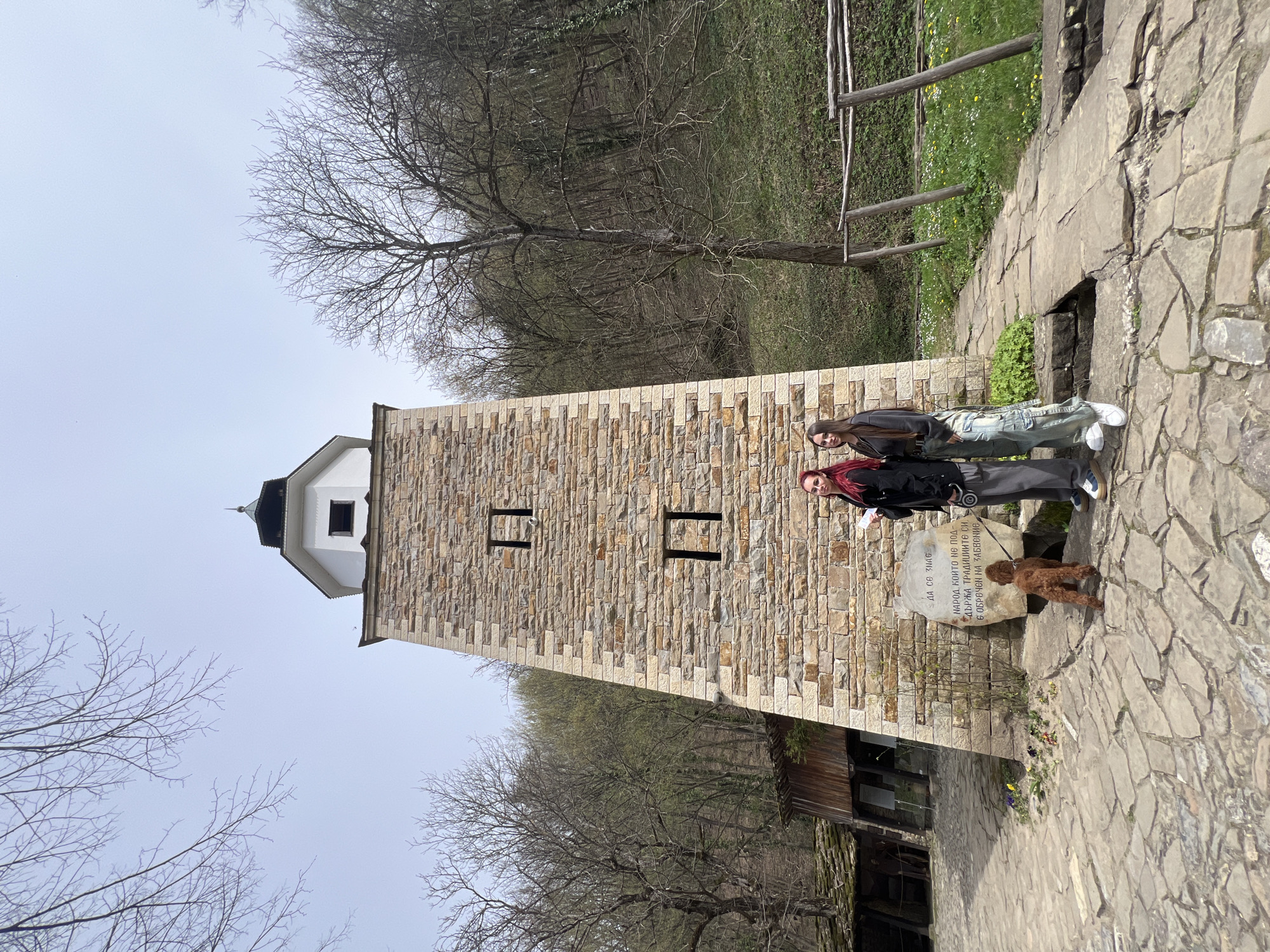 Here's a detailed description of the image:
**General Description:**
The image shows a vertical view of a stone tower and surrounding area. Two people are standing on a stone pathway next to the tower, and a small dog is nearby. The overall setting appears to be outdoors, possibly a historical site or park.
**Environment and Objects:**
* **Stone Tower:** A tall, rectangular tower constructed from light-colored stone bricks dominates the scene. The tower has two small, dark rectangular openings in the middle. The top portion is painted white with a black, roof.
* **Stone Pathway:** The ground consists of a stone pathway constructed from irregular stone slabs.
* **Stone Monument:** A stone monument stands next to the walkway, appearing next to the tower with writing in Bulgarian.
**People:**
* Two people are standing on the stone pathway beside the tower.
* **Dog:** A small, brown dog is standing on the pathway in front of the tower and next to the rock monument.
**Location:**
Based on the visual information, the image depicts the tower in Gabrovo, Bulgaria.