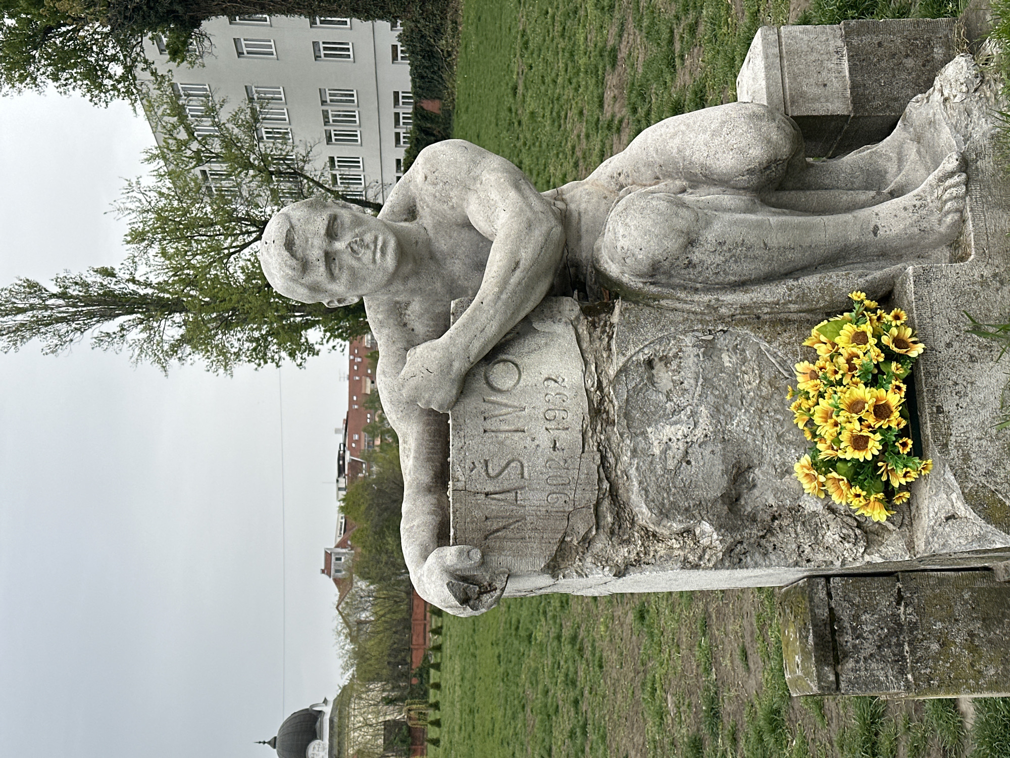 The image depicts a horizontal, eye-level view of a weathered stone memorial in Novi Sad, Serbia. The statue features a reclining male figure with a solemn expression, resting upon a stone surface bearing the inscription "NAS IVO 1902-1932". A cluster of yellow flowers sits next to the figure. The surrounding environment is a grassy park, with buildings, trees, and a dome structure visible in the background. The sky is overcast.
